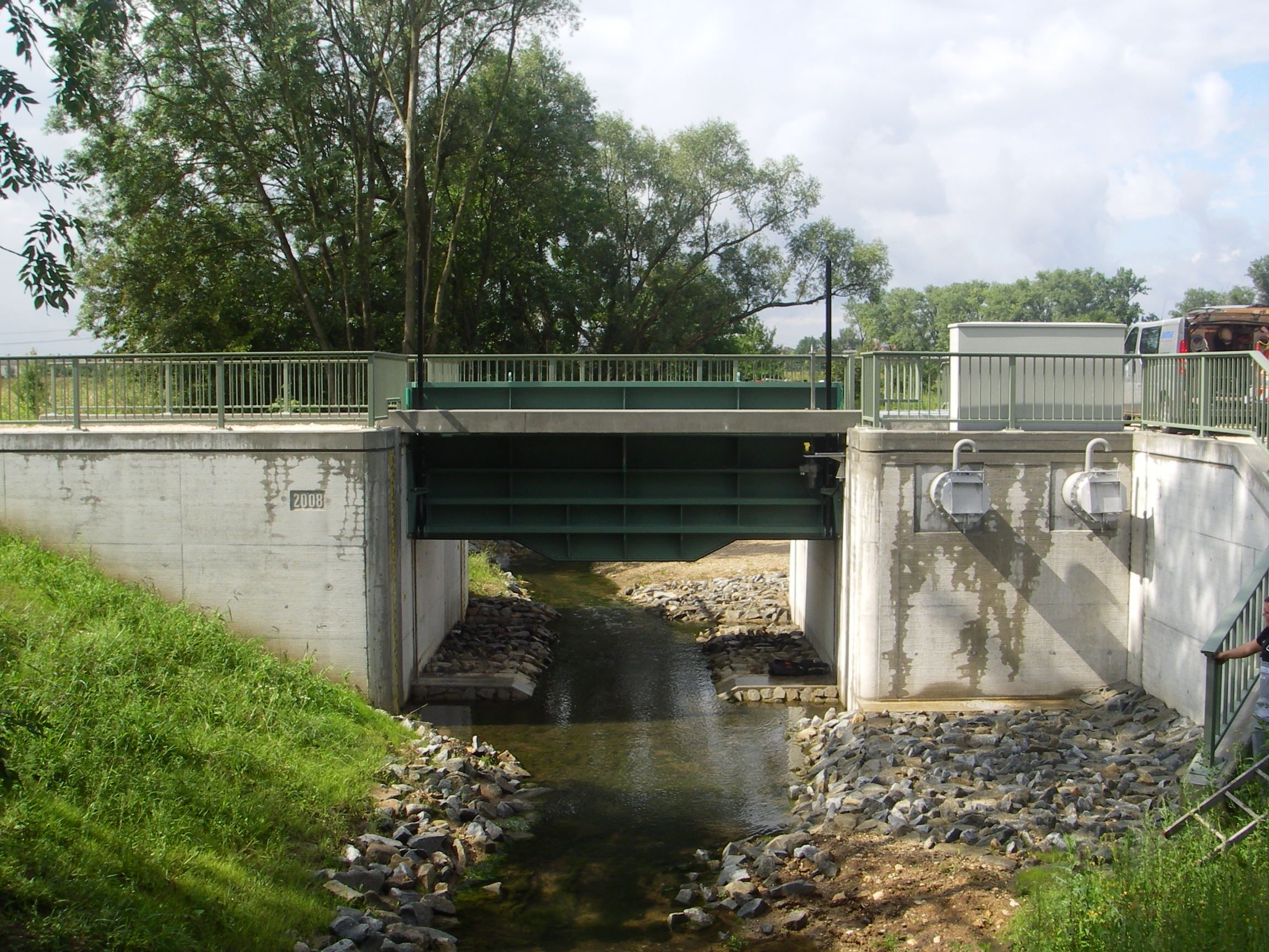 In der Mitte des Bildes nach hinten verlaufend der Aubach. Quer darüber das betonierte Schöpfwerk mit Schütztafel und rechts die beiden Rohrschächte, aus denen das Wasser im Hochwasserfall gepumpt wird. Vor diesen Schächten ist der Boden gepflastert. Auf dem Übergang und rechts die Treppe hinunter zum Aubach ist ein grünes Geländer angebracht. Links vor dem Schöpfwerk ist die Böschung begrünt, im Hintergrund stehen Bäume.