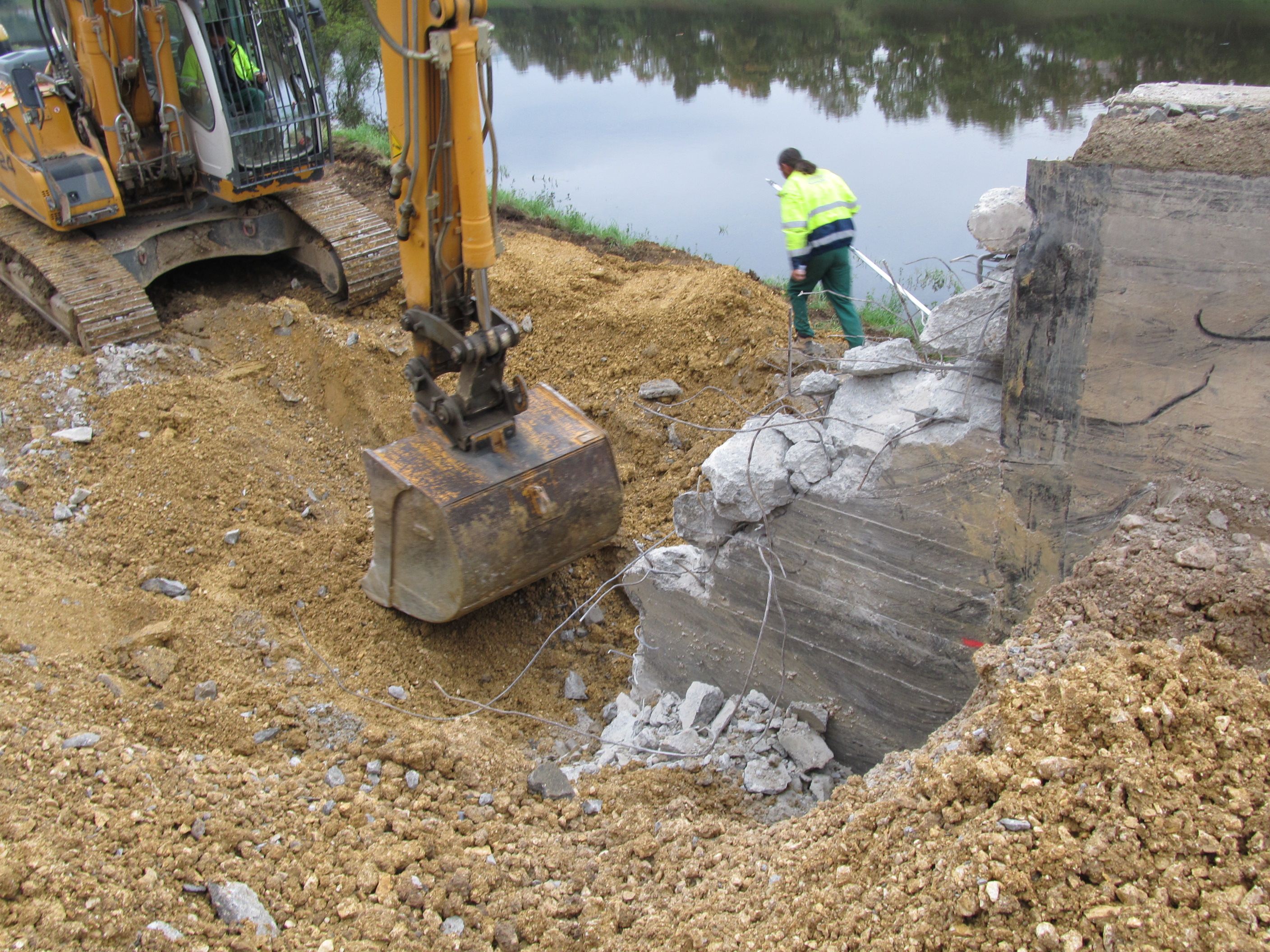 Links im Bild ein gelber Bagger auf sandigem Untergrund. Mit einer Schaufel bricht dieser eine rechts im Bild befindliche Betonwand mit Baustahl ab. Im Hintergrund der Fluss Regen, dazwischen ein Bauarbeiter mit neongelber Arbeitsjacke