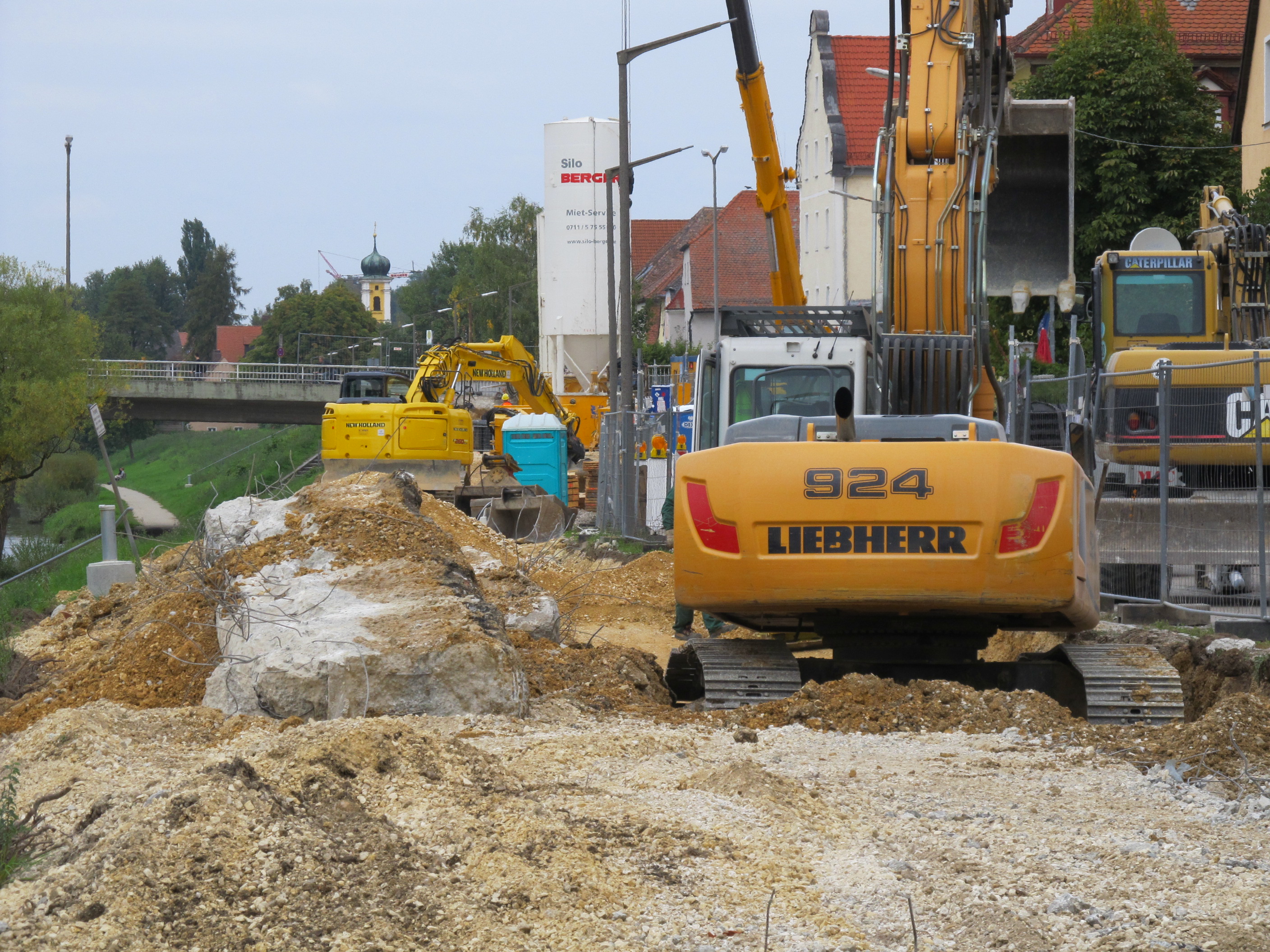 Auf dem unebenen, sandigen Gelände befinden sich von links eine Grundwassermessstelle, abgebrochene Betonstücke, zwei gelbe Bagger, ein Betonsilo, mobile Toilette, Bauzäune und Absperrungen. Rechts im Bild ist die Bebauung der Unteren Regenstraße zu erkennen. Im Hintergrund quert die Reinhausener Brücke und die Turmspitze von St. Nikolaus ist zu sehen.