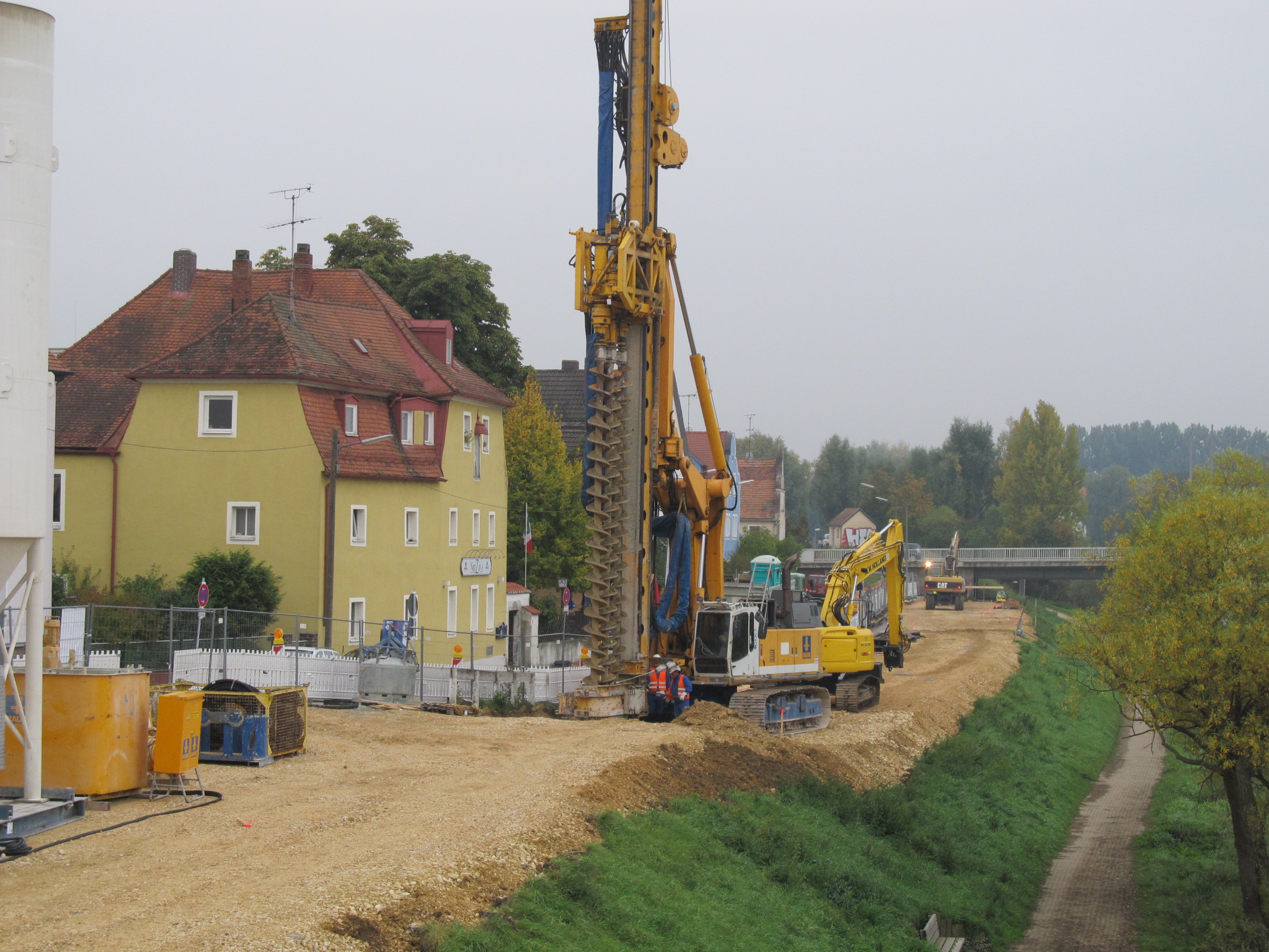 In der Mitte des Bildes steht das Bohrgerät, zwei Bauarbeiter und ein Bagger auf der planierten Hochwasserschutztrasse. Links davon ein Bauzaun, die Bebauung und Teile der Baustelleneinrichtung. Dahinter fährt ein Bagger auf die Frankenbrücke zu. Rechts im Bild die Böschung und der Uferweg.