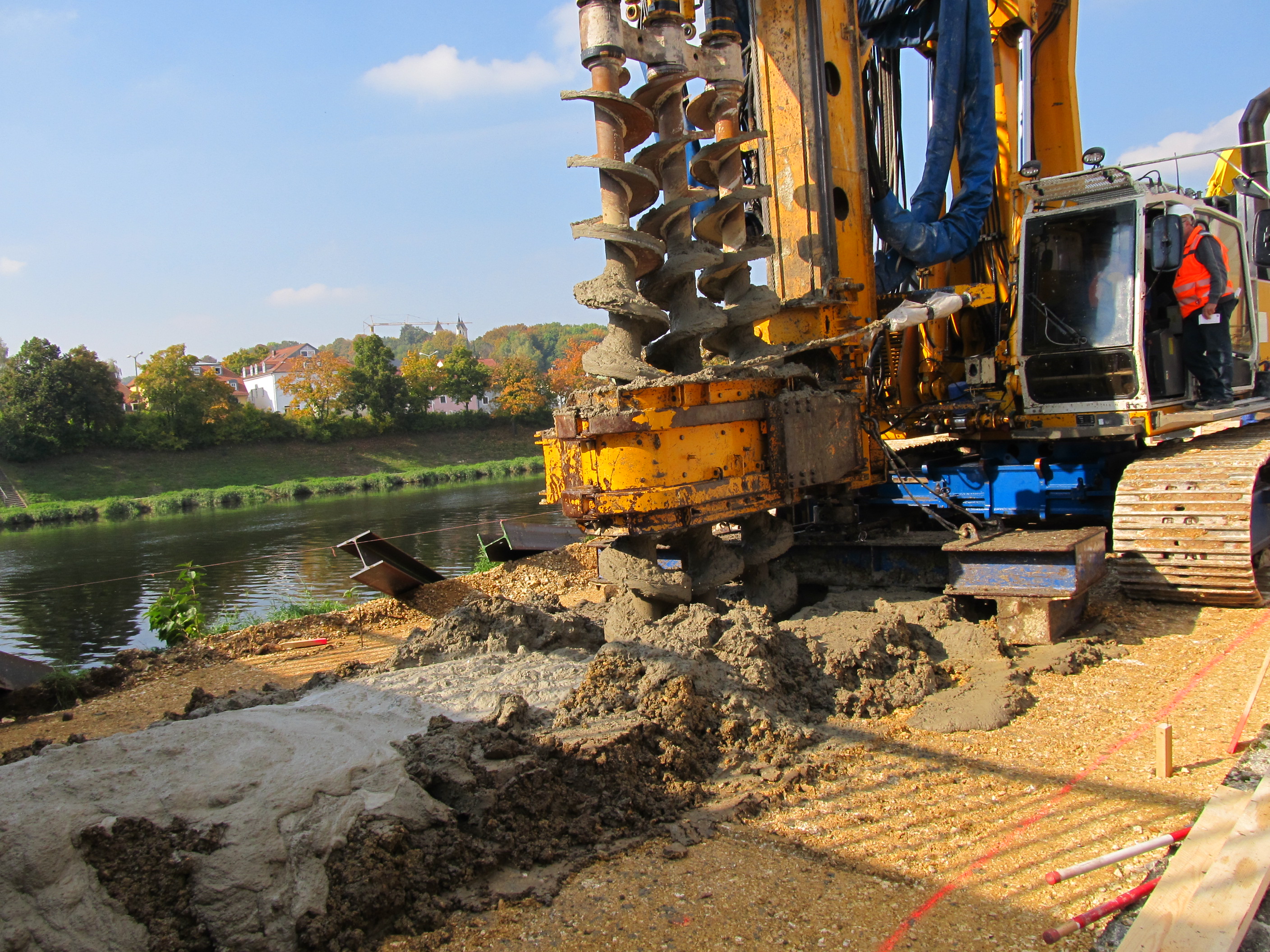 Rechts im Bild steht das Bohrgerät. Erdreich wird entnommen und Beton eingebracht, in einem Arbeitsgang. Dieses Verfahren wird MIP, mixed in place genannt. Im Hintergrund ist der Fluss Regen und das gegenüberliegende Ufer mit Böschung, Bepflanzung und Bebauung zu sehen.