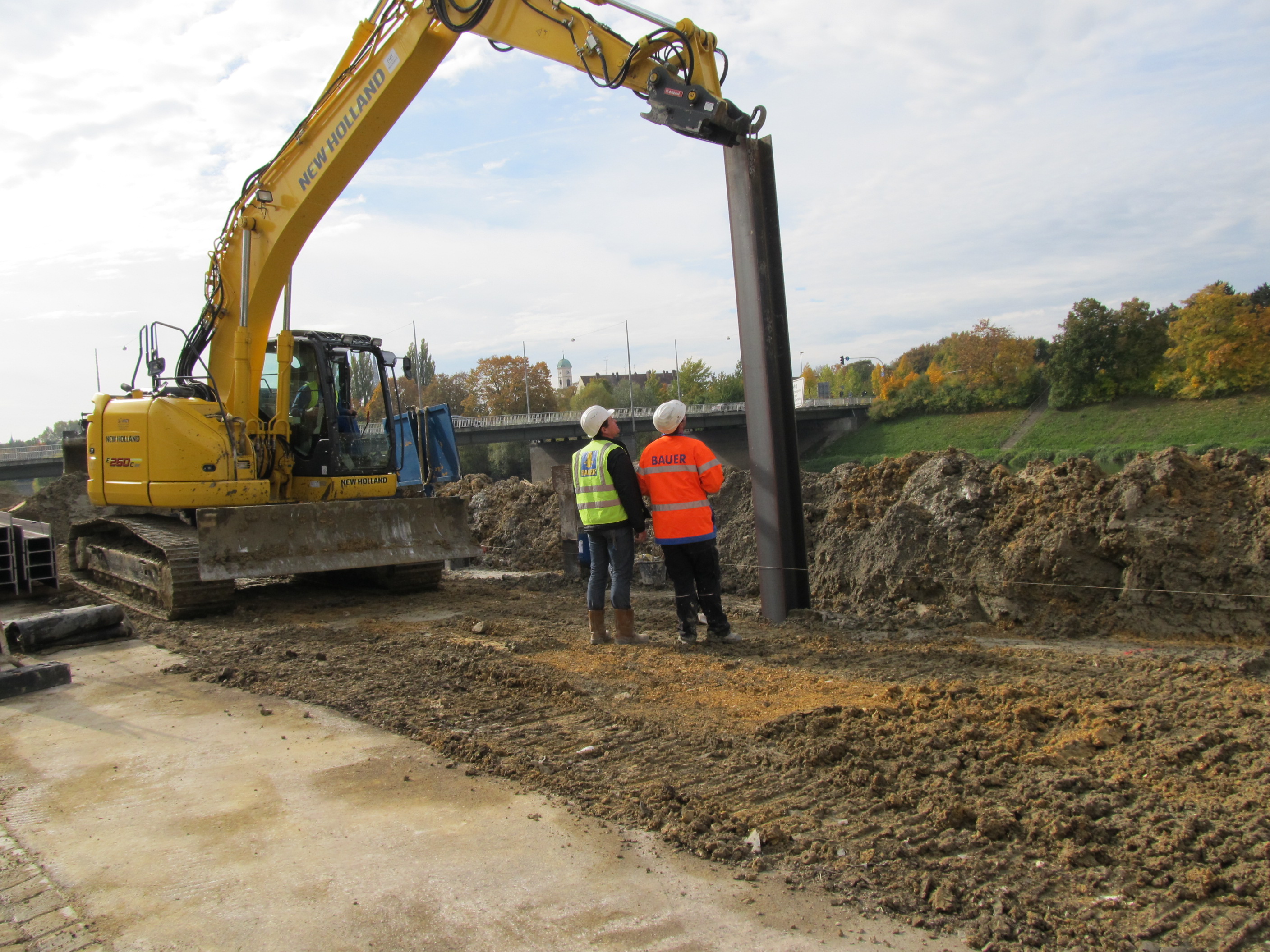 Links im Bild ein Bagger, der einen Stahlträger in die MIP, mixed in place, Wand in der Mitte des Bildes einbringt. Zwischen Bagger und Stahlträger verfolgen zwei Bauarbeiter den Arbeitsschritt. Im Hintergrund liegt Aushubmaterial, dahinter quert die Frankenbrücke den Fluss Regen. Die gegenüberliegende Böschung und das Ufer ist zu sehen und der Turm der St. Mang Kirche in Stadt am Hof.