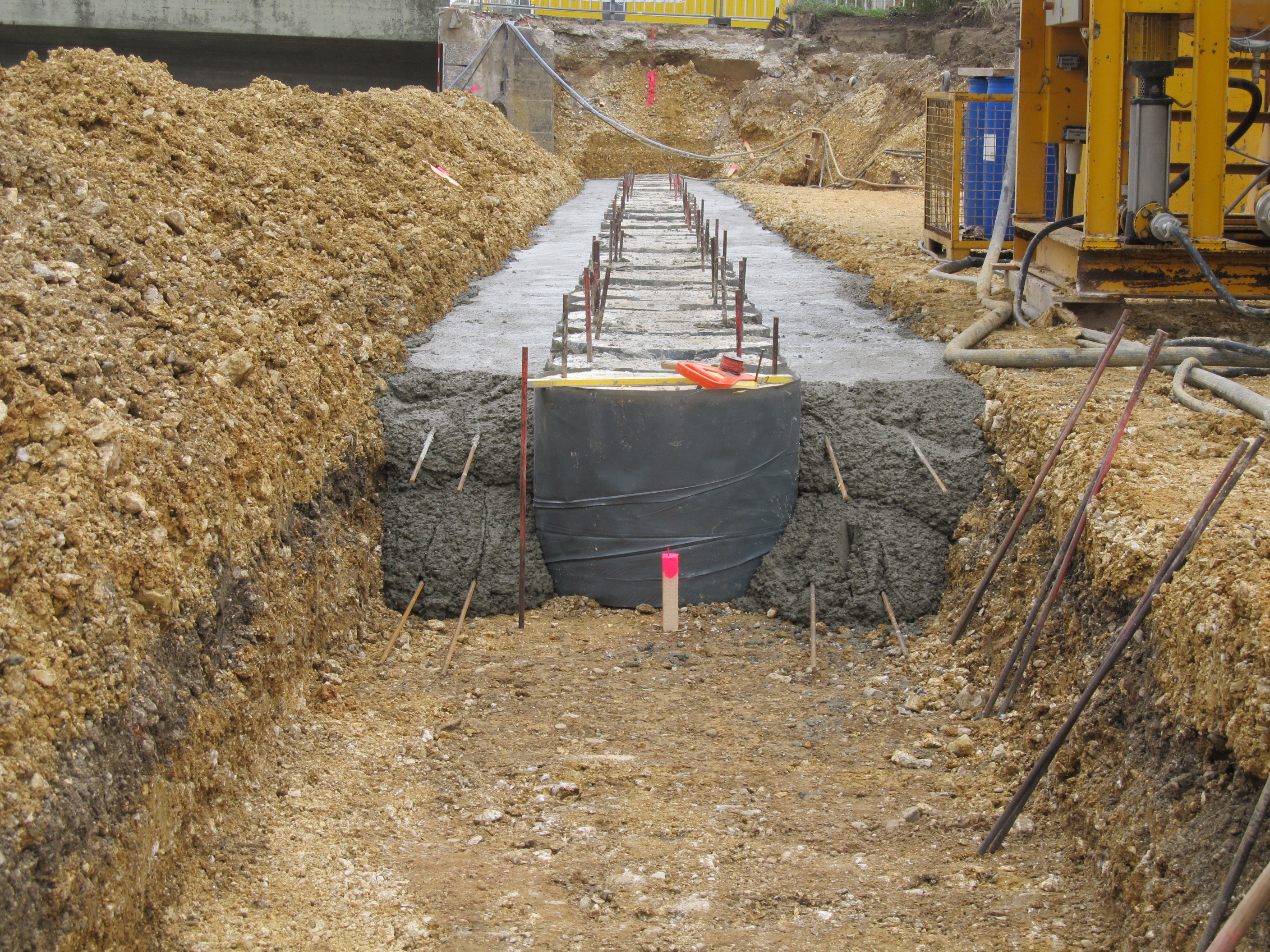 In Bildmitte nach hinten verlaufend werden in einer Grube Styroporschablonen eingebracht und an beiden Seiten bis zum Grubenrand mit Baustahl versehener Beton aufgefüllt. Links liegt das ausgehobene Material, rechts stehen Baugeräte. Im Hintergrund ist das Widerlager der Reinhausener Brücke zu erkennen.