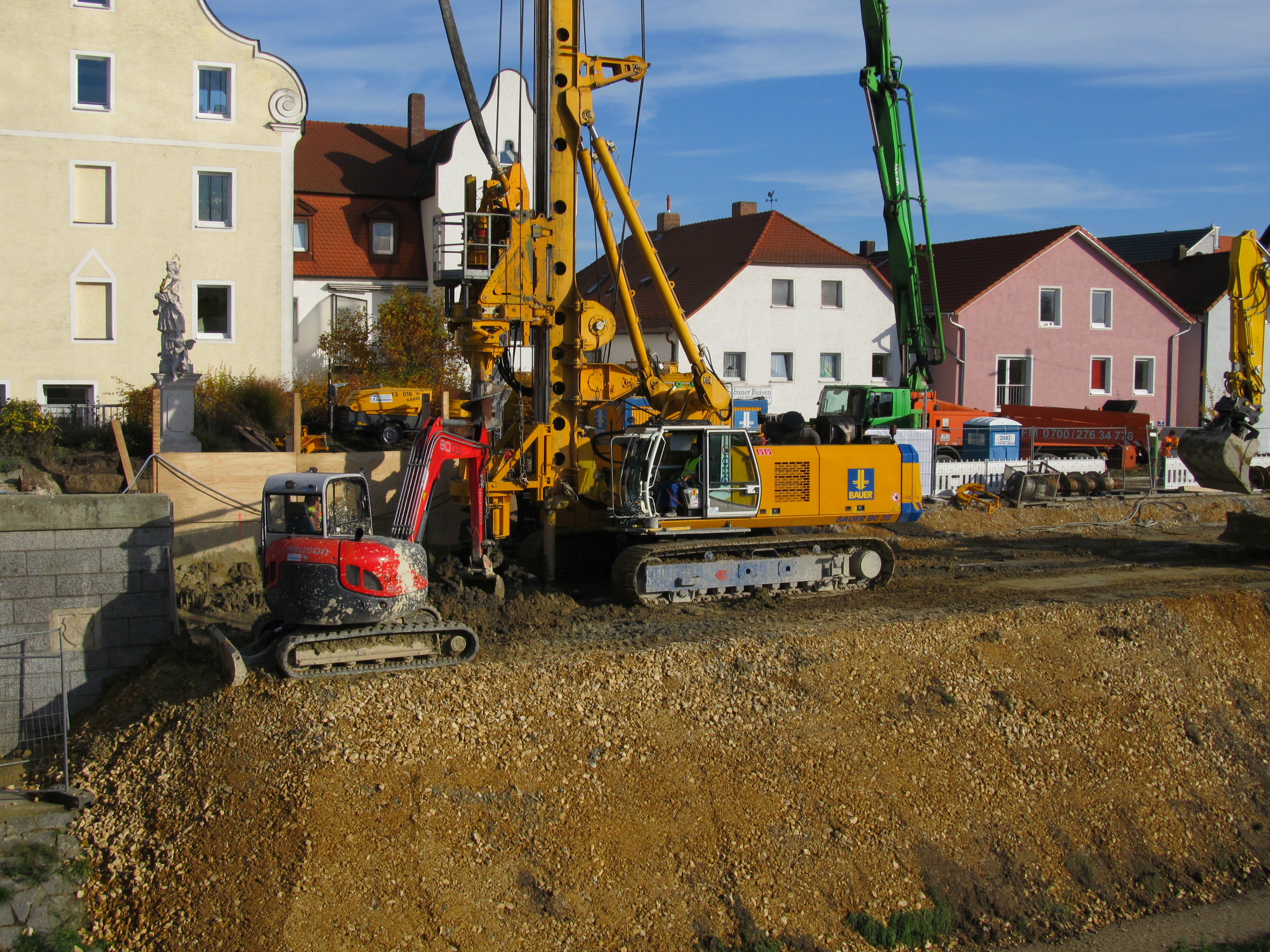 Auf der mit Schotter aufgefüllten Böschung arbeitet das Bohrgerät, links davon ein kleiner Bagger, der den Aushub beseitigt. Im Hintergrund Baumaschinen und Bauutensilien. Dahinter die Häuserreihe der Unteren Regenstraße.