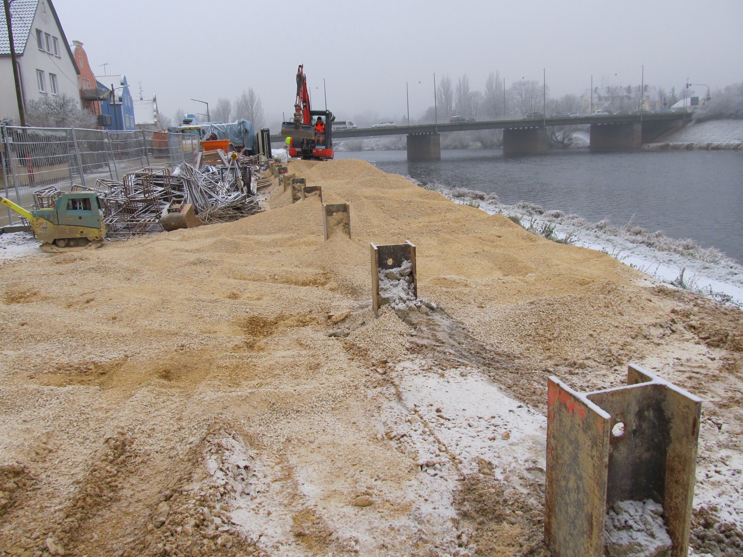 In der Mitte des Bildes, nach hinten verlaufend stehen Stahlträgerenden zum Anschluss der Hochwasserschutzmauer aus der MIP, mixed in place, Wand heraus. Im Hintergrund ein Bagger, dieser füllt das Gelände mit Schotter auf. Links davon Bauutensilien und Baumaschinen, der Bauzaun, die Straße und die Häuserreihe. Rechts davon der Fluss Regen über den die Frankenbrücke quert.