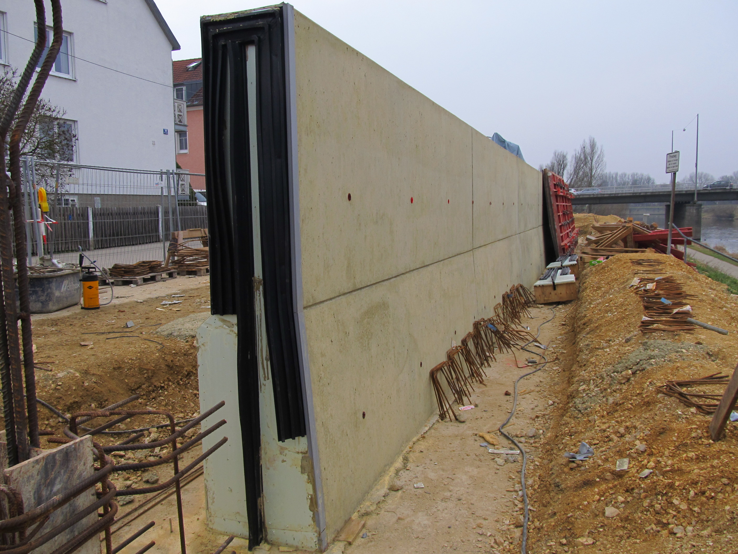 Leicht schräg in der Mitte des Bildes steht die Hochwasserschutzwand, welche zum Teil schon ausgeschalt ist. Am hinteren Ende ist die Schalung noch vorhanden. Links im Bild die Straße und die Häuserreihe, rechts der Fluss Regen und die Frankenbrücke im Hintergrund.