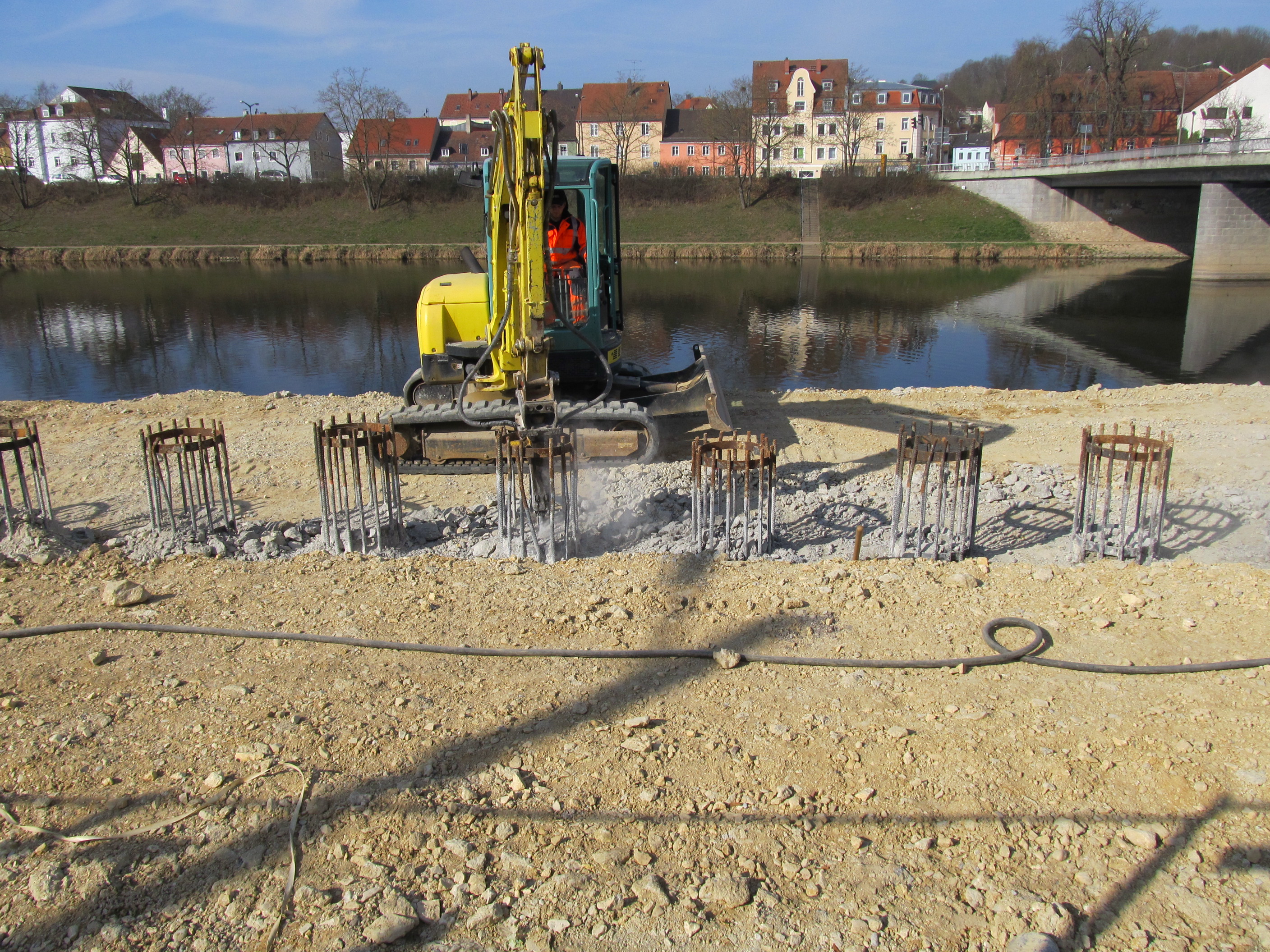 In der Mitte des Bildes steht eine Baumaschine, die mit einem Meisel den überständigen Beton der sieben Bohrpfähle aufstemmt. Die Bewehrung der Bohrpfahlwand steht gut einen halben Meter über die Geländeoberkante heraus. Im Hintergrund quert der Fluss Regen, dahinter ist das Ufer und die Bebauung zu sehen. Rechts im Bild steht die Reinhausener Brücke.