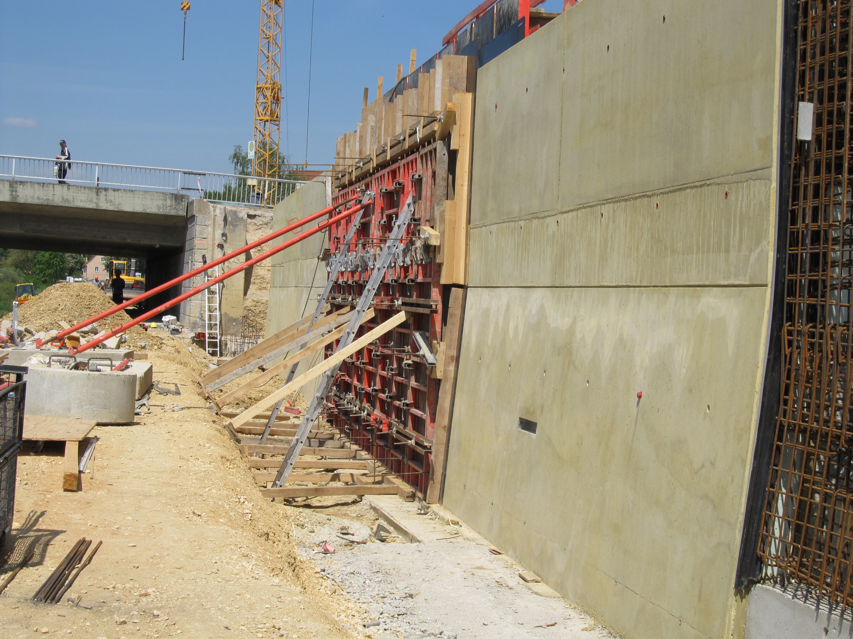 Rechts im Bild die Hochwasserschutzwand. Teilweise geschalt, betoniert oder nur mit Baustahl. Links der zukünftige Unterhaltungsweg mit Baumaterial, im Hintergrund die Reinhausener Brücke und ein gelber Baukran. Unter der Brücke hindurch sind Bauarbeiter und Baumaschinen zu erkennen. Ein Passant steht auf der Brücke.