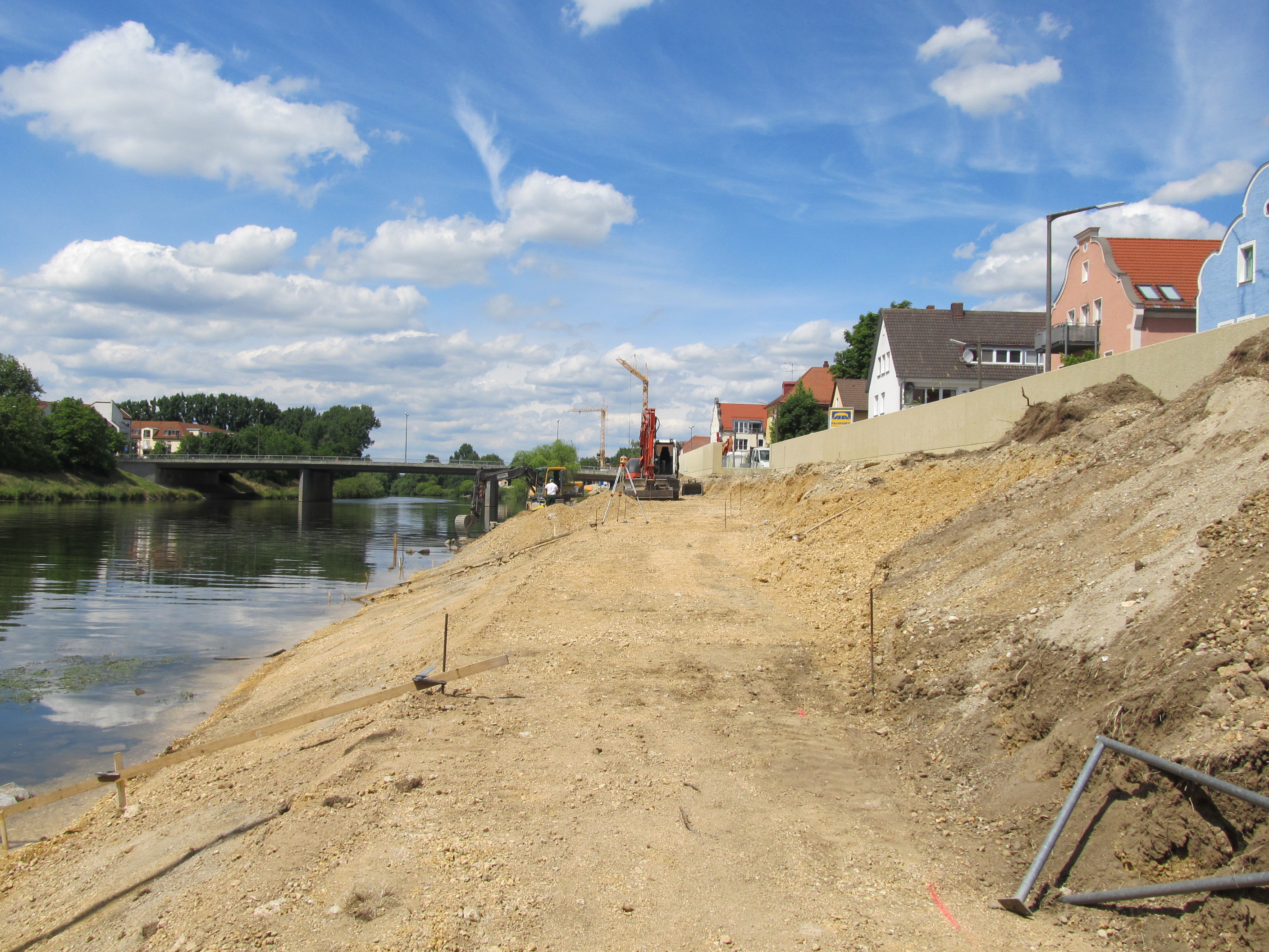 Rechts im Bild wird der Unterhaltungsweg mit einem Bagger angelegt und abgezogen. Rechts die Böschung, die Hochwasserschutzwand und die Häuserreihe. Links der Fluss Regen, das linke Regenufer mit Bewuchs. Im Hintergrund quert die Reinhausener Brücke und ein Wohnblock ist zu sehen, bei leicht bewölktem Himmel.