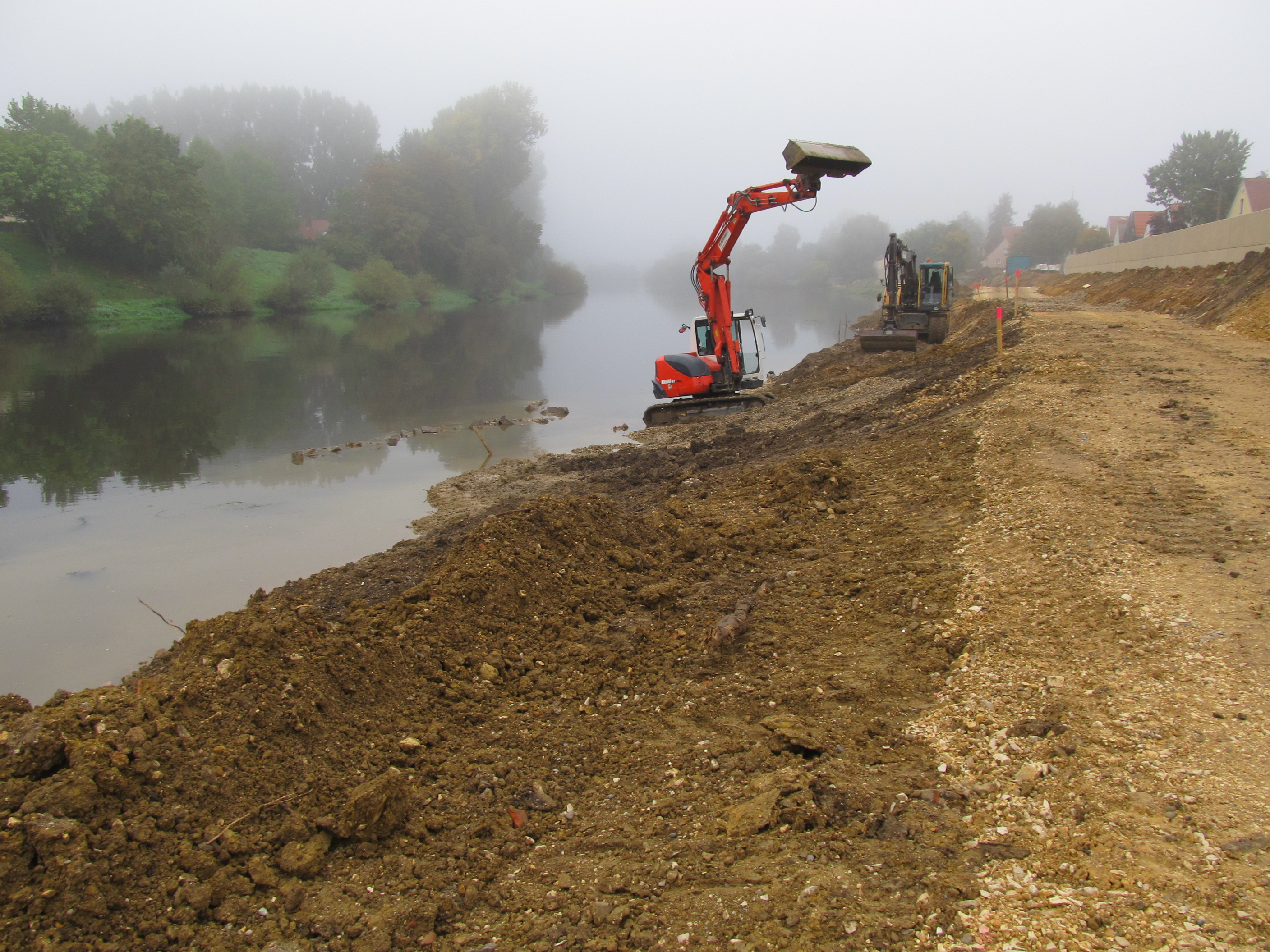  An der rechten Bildseite arbeiten zwei Bagger an der Geländemodellierung am Regenufer, links im Bild der Fluss Regen und das linke Regenufer mit Bewuchs im Frühnebel.