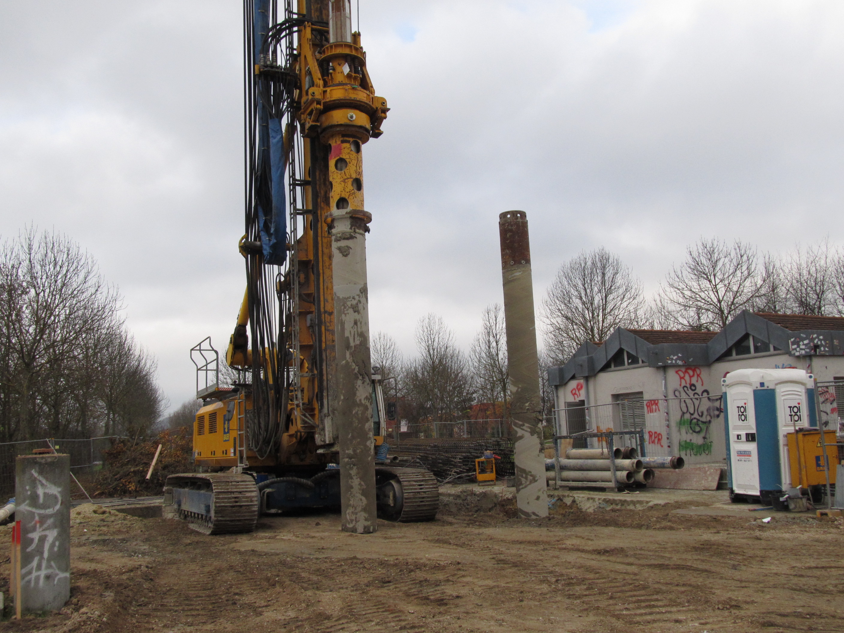 In der Mitte des Bildes das Bohrgerät auf unbefestigtem Grund, rechts im Bild der mit Graffiti besprühte Hochbau einer Pumpstation, Bauutensilien und eine mobile Toilette.