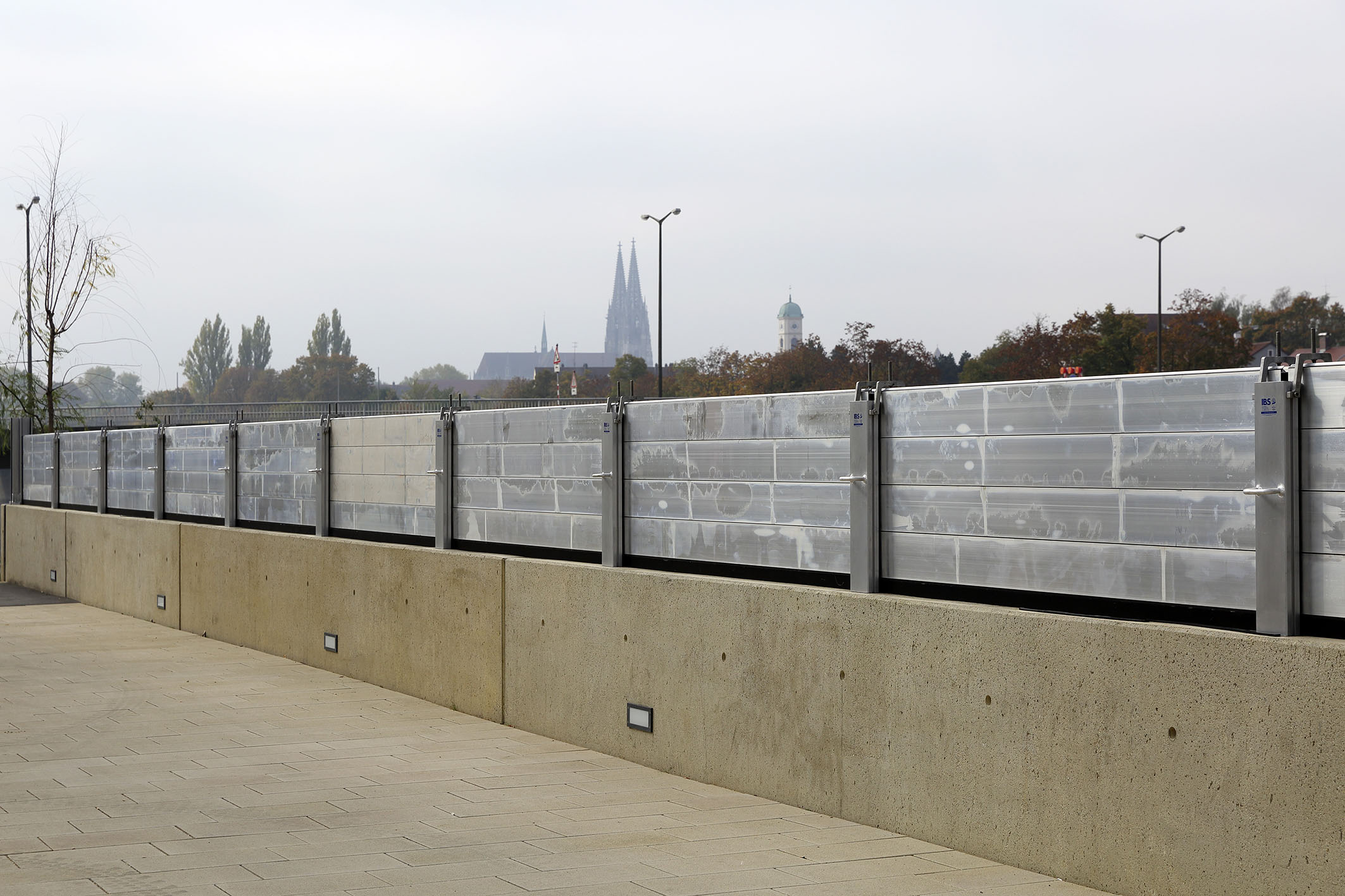 Diagonal im Bild verläuft die Hochwasserschutzwand mit den aufgesetzten mobilen Elementen.  Im Hintergrund ist die Bebauung, das Geländer und die Laternen der querenden Reinhausener Brücke, der Dom und die Turmspitze der St. Mang Kirche in Stadt am Hof zu sehen.