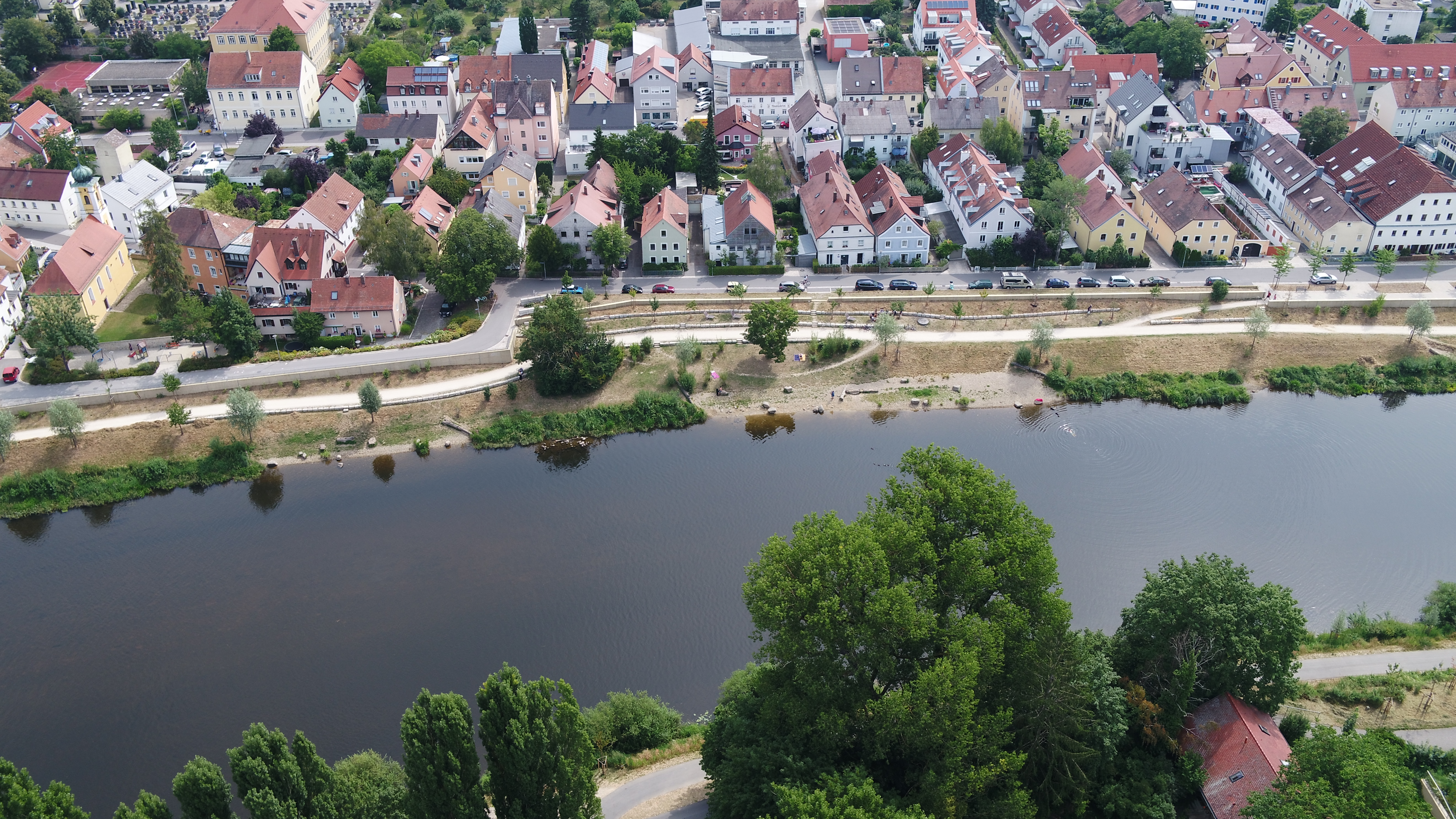 Horizontal durch das Bild fließt der Fluss Regen. Im oberen Teil des Bildes die bereits eingewachsene Hochwasserschutzmaßnahme. Dazu zählen die Hochwasserschutzwand entlang der Böschung, ein neuer Geh- und Fahrradweg, Flachuferbuchten, neue Bepflanzung und Sitzgelegenheiten. Dahinter die neu angelegte Straße, ein kleiner Spielplatz und Bebauung. Am gegenüberliegenden Ufer ist der Radweg zu sehen mit Bäumen und einem Hausdach.