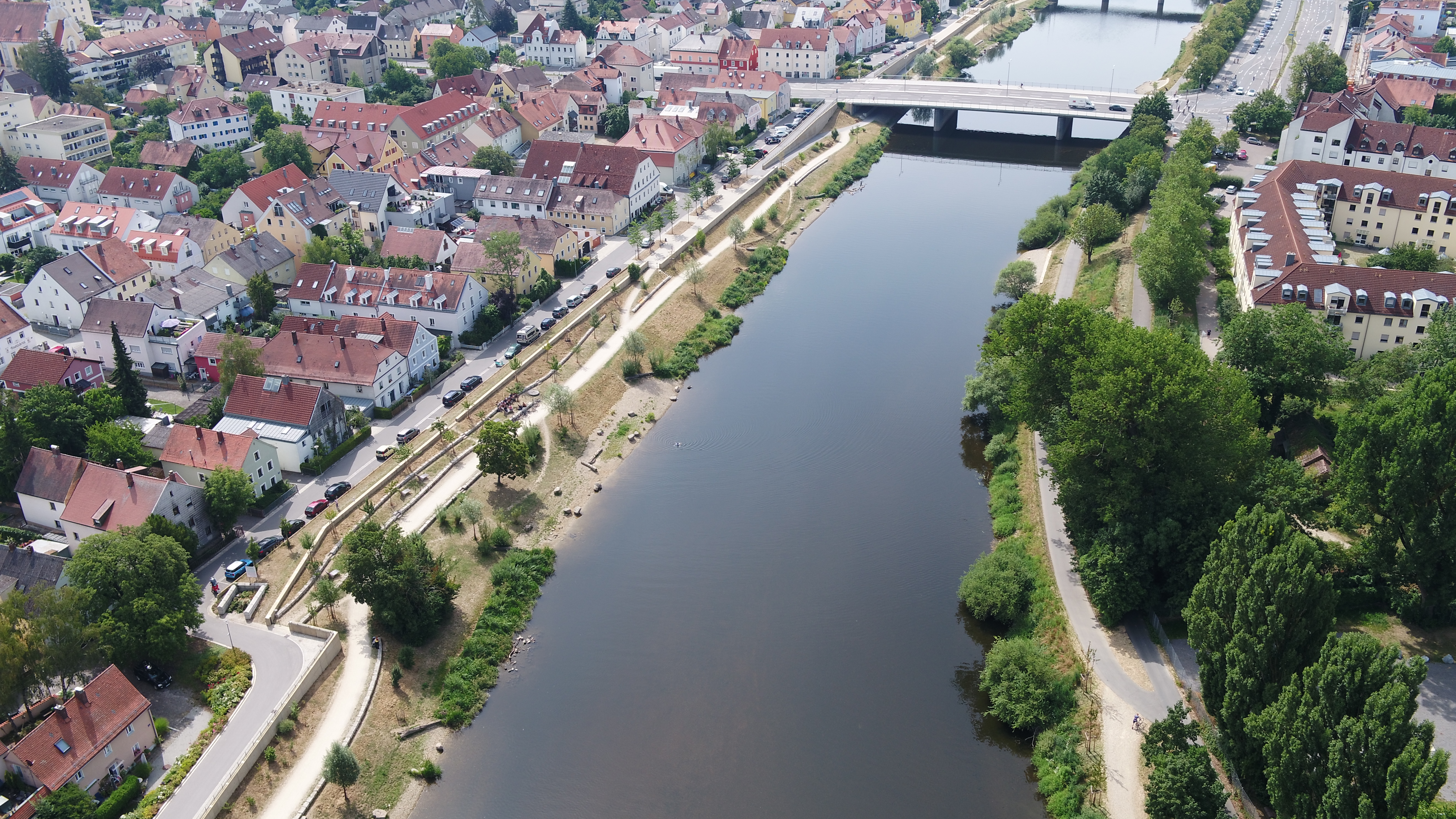 In der Mitte des Bildes der Fluss Regen mit einer Brücke im Hintergrund, am linken Regenufer die bereits eingewachsene Hochwasserschutzmaßnahme. Dazu zählen die Hochwasserschutzwand entlang der Böschung, ein neuer Geh- und Fahrradweg, Flachuferbuchten, neue Bepflanzung und Sitzgelegenheiten. Dahinter die neu angelegte Straße und Bebauung. Am rechten Ufer die Böschung, der Radweg und die Bebauung