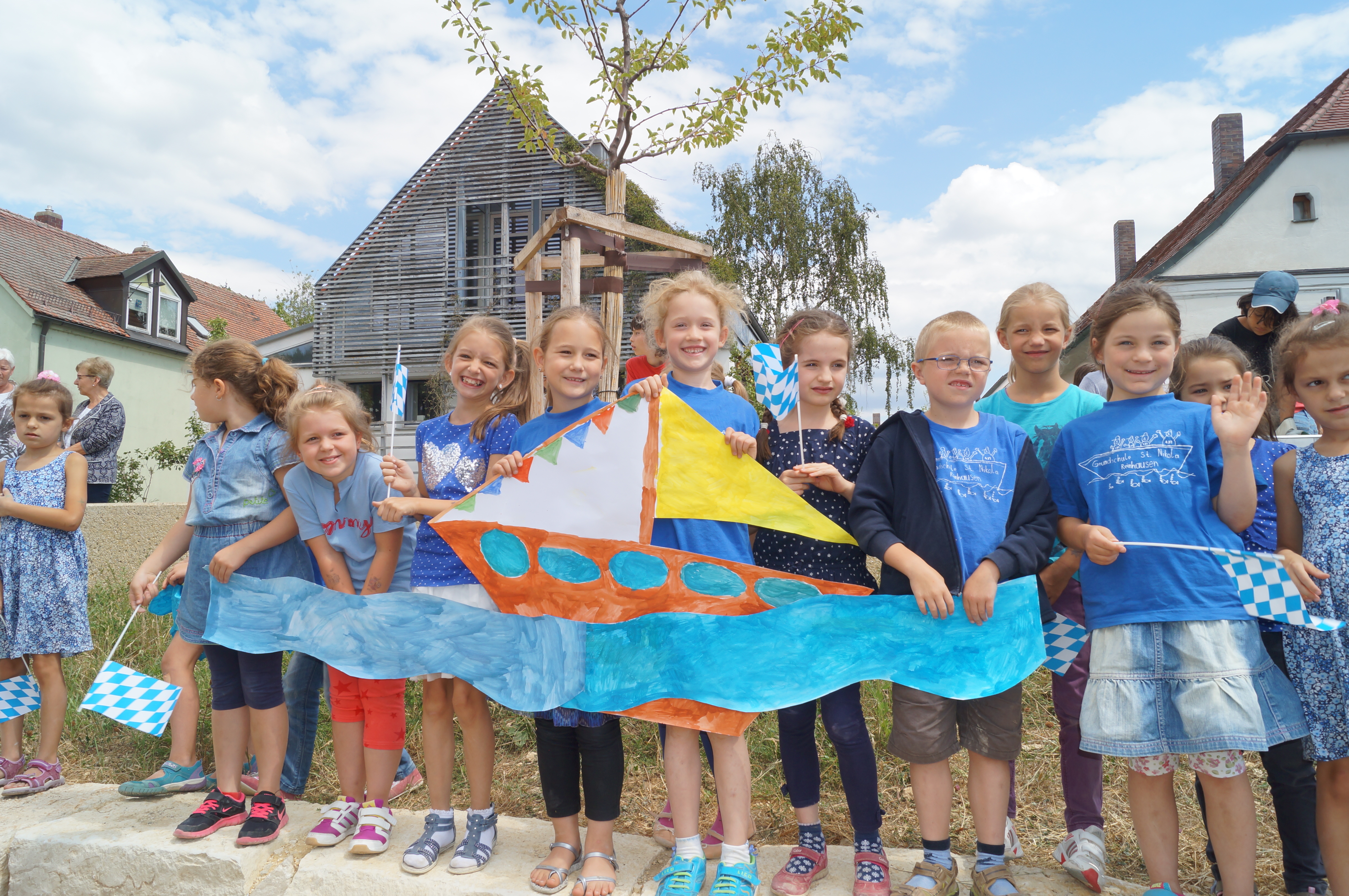 Fröhliche Kinder der Grundschule halten ein aus Papier ausgeschnittenes und bemaltes Schiff mit dem ausgeschnittenen und bemalten Fluss Regen vor sich. Einige der Kinder winken mit kleinen Bayernfähnchen. Im Hintergrund Bäume und Bebauung.