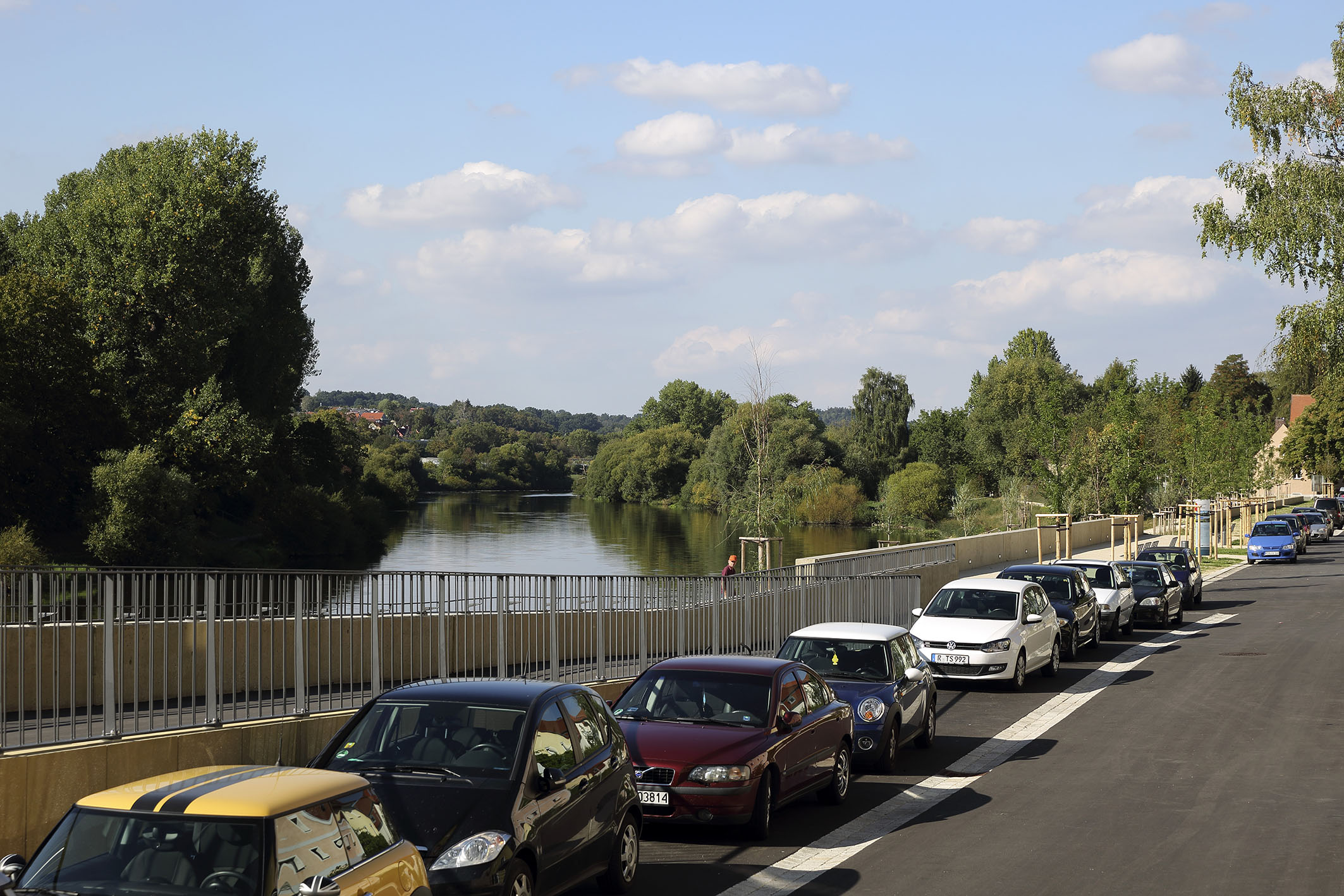  Im Hintergrund der Fluss Regen mit beidseitigem Uferbewuchs, im Vordergrund quert die Obere Regenstraße mit der Auffahrt für Radfahrer und Fußgänger zur Reinhausener Brücke mit beidseitigem Geländer. Im weiteren Verlauf steht die Hochwasserschutzmauer mit neu gepflanzten Bäumen. Davor eine Reihe parkender Autos entlang der Straße bei leicht bewölktem Himmel.