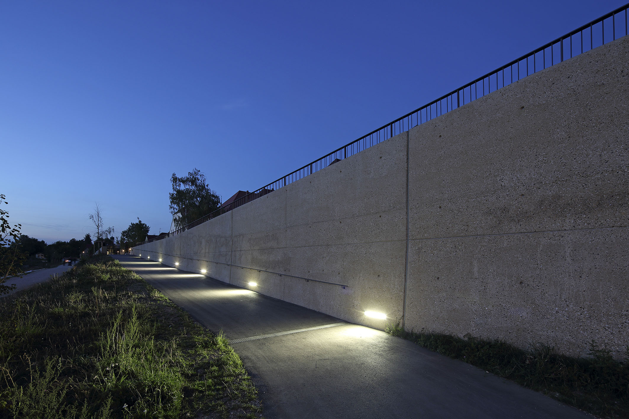 In der Mitte des Bildes der asphaltierte Radweg, rechts davon die Hoschwasserschutzmauer mit aufgesetztem Geländer und bodennaher Beleuchtung bei Nacht, links davon die Böschung und der Unterhaltungsweg