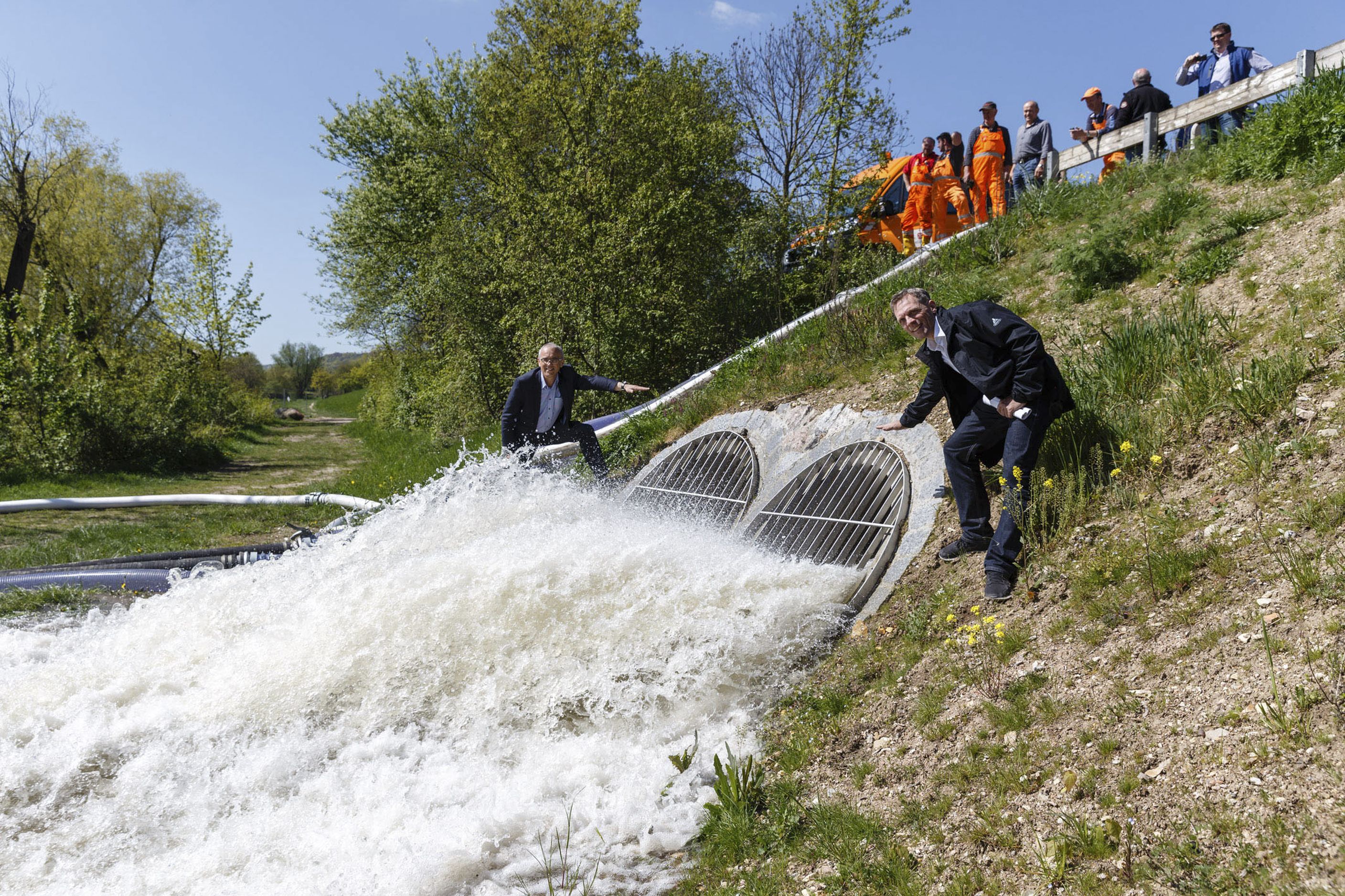 In der Mitte des Bildes sind die Öffnungen das Pumpwerks mit ausströhmenden Wasser zu sehen. Rechts davon steht Hr. Zimmermann, Projektleiter für den Hochwasserschutz in Regensburg vom Wasserwirtschaftsamt, links davon Hr. Kastenmeier vom Tiefbauamt der Stadt Regensburg. Im Hintergrund liegen Wasserschläuche und die Bepflanzung ist zu sehen. Rechts oberhalb der begrünten Böschung stehen Mitarbeiter des Wasserwirtschaftsamtes Regensburg.