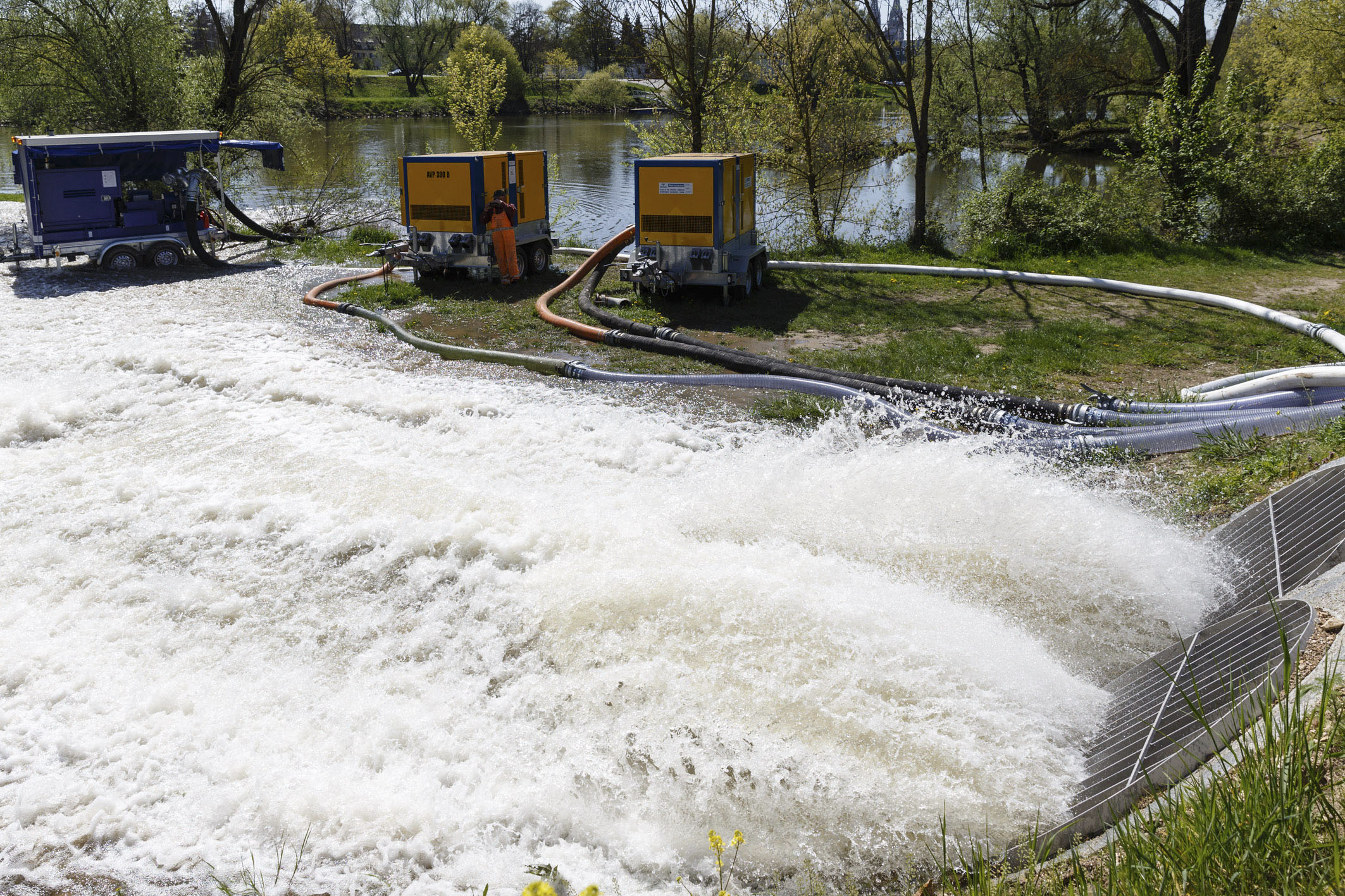 Im Hintergrund quert die Donau mit Uferbewuchs. Davor ströhmt Wasser aus dem rechts im Bild zu sehenden Öffnungen des Pumpwerks. In der Mitte des Bildes stehen drei zusätzliche mobile Pumpen mit Schläuchen und ein Bauarbeiter fotografiert das Geschehen.