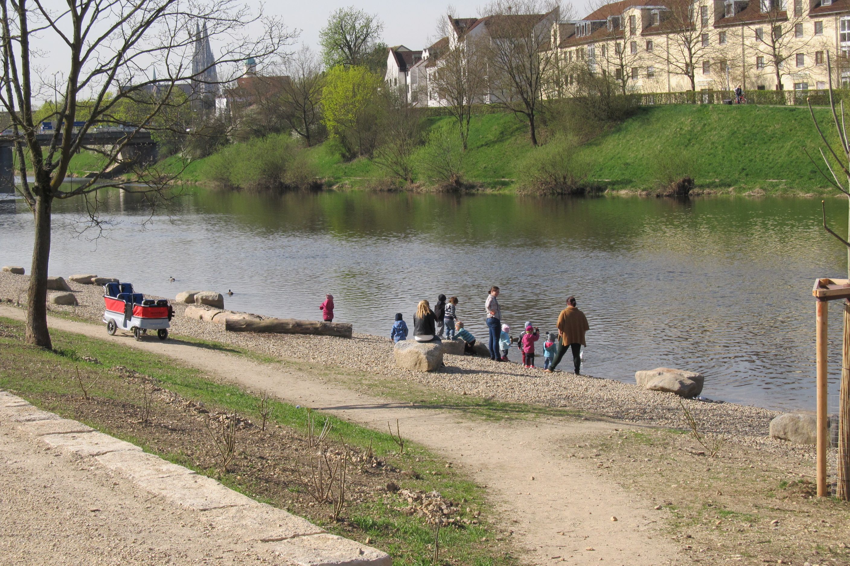 Quer durch das Bild der Fluss Regen mit darin schwimmenden Enten. Im Vordergrund der neu angelegte Kieselstrand mit Erwachsenen und Kindern. Am Uferweg steht ein Bollerwagen. Uferbewuchs und Bebauung am gegenüberliegenden Ufer. Im Hintergrund ist die Frankenbrücke, der Dom und der Kirchturm von St. Mang zu sehen.