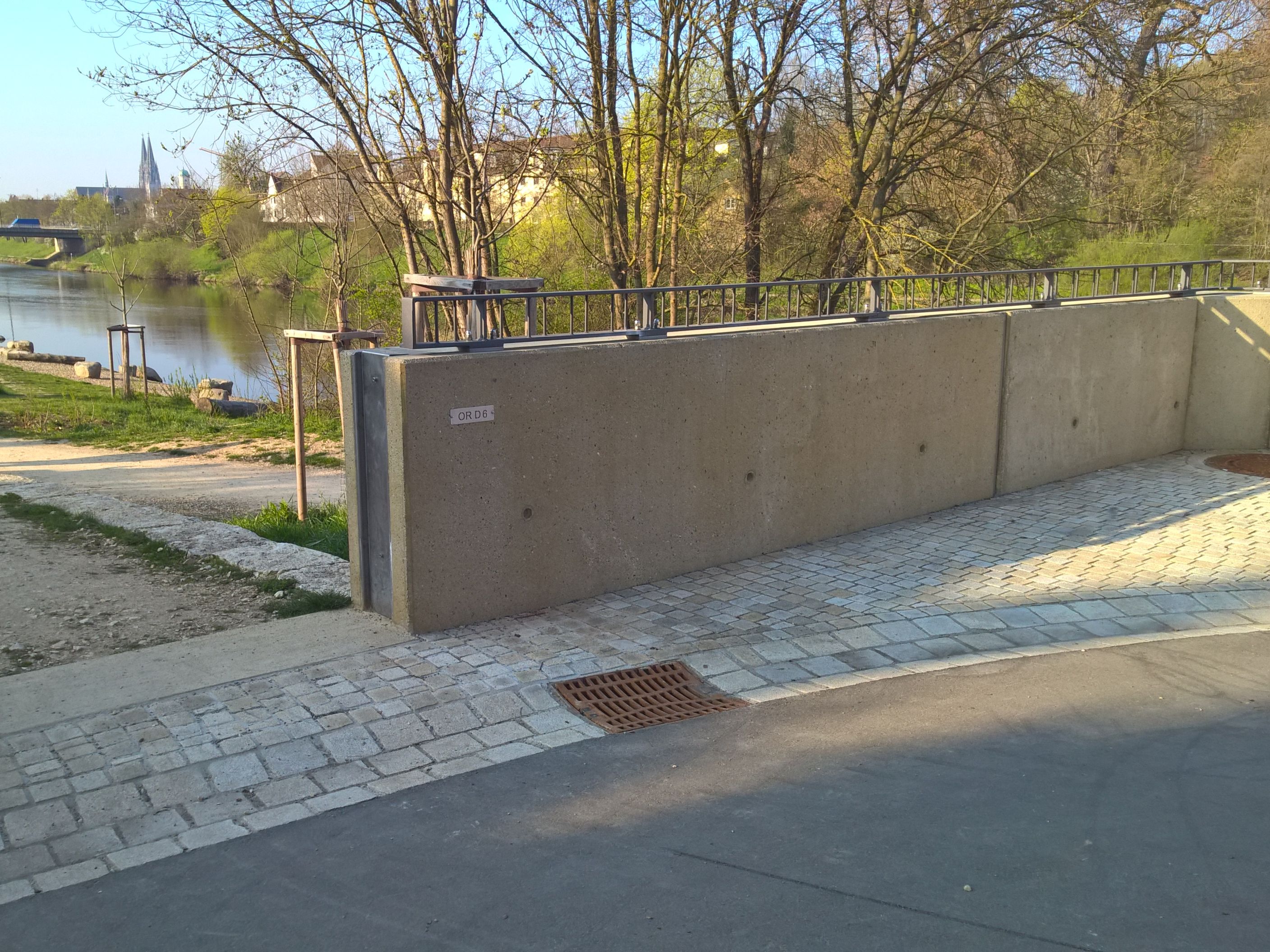 Rechts und quer im Bild die Hochwasserschutzmauer mit aufgesetztem Geländer in der Uferstraße. Von links nach rechts sind der Uferweg mit Mülleimer, die Bepflanzung, das Kiesufer mit Steinen und Baumstämmen, der Fluß Regen und das linke Regenufer mit Bepflanzung und Bebauung zu sehen. Im Hintergrund quert die Reinhausener Brücke und der Dom ist zu sehen.