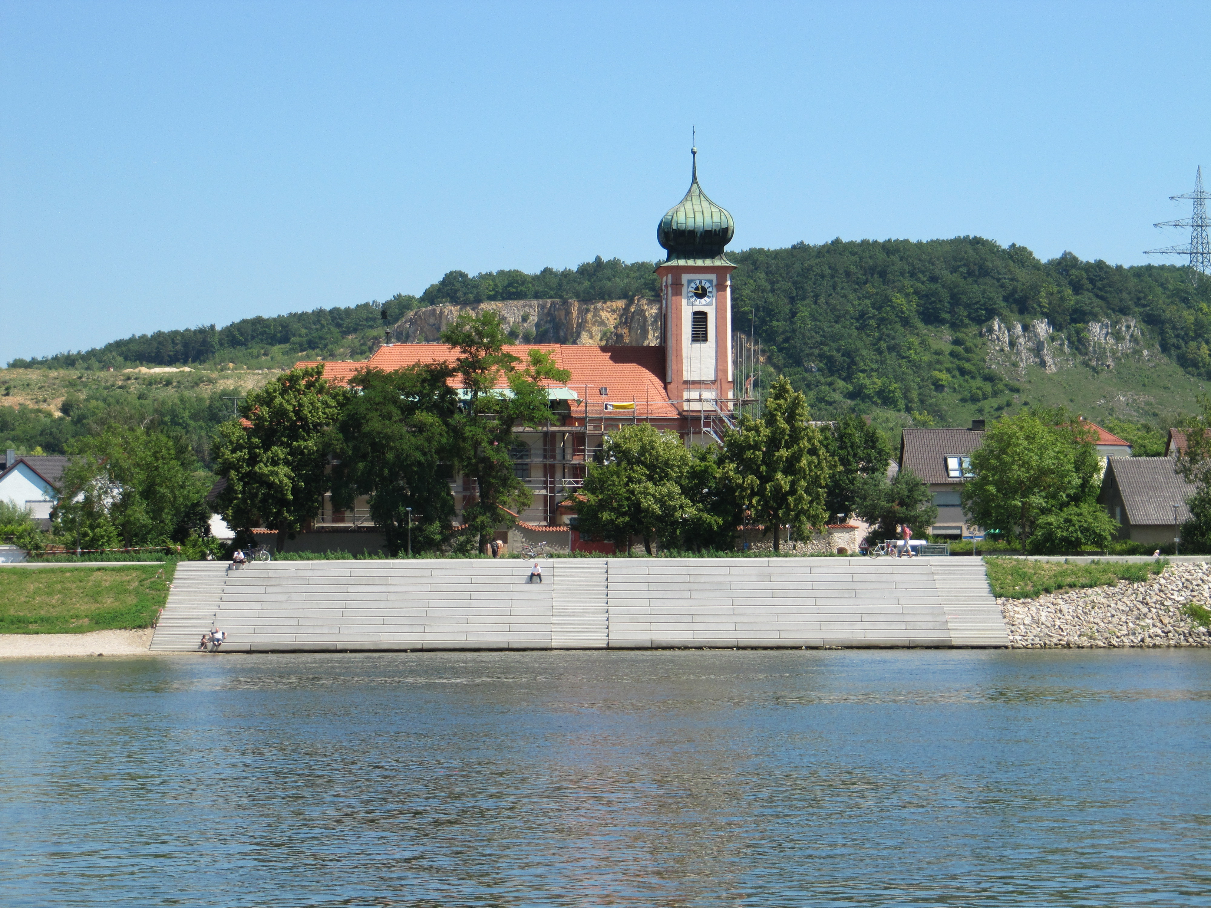 Die Donau im Vordergrund, dahinter die Treppenanlage aus Beton mit vier darauf sitzenden Personen. Im Hintergrund eine Laubbaumreihe und die eingerüstete Schwabelweiser Kirche