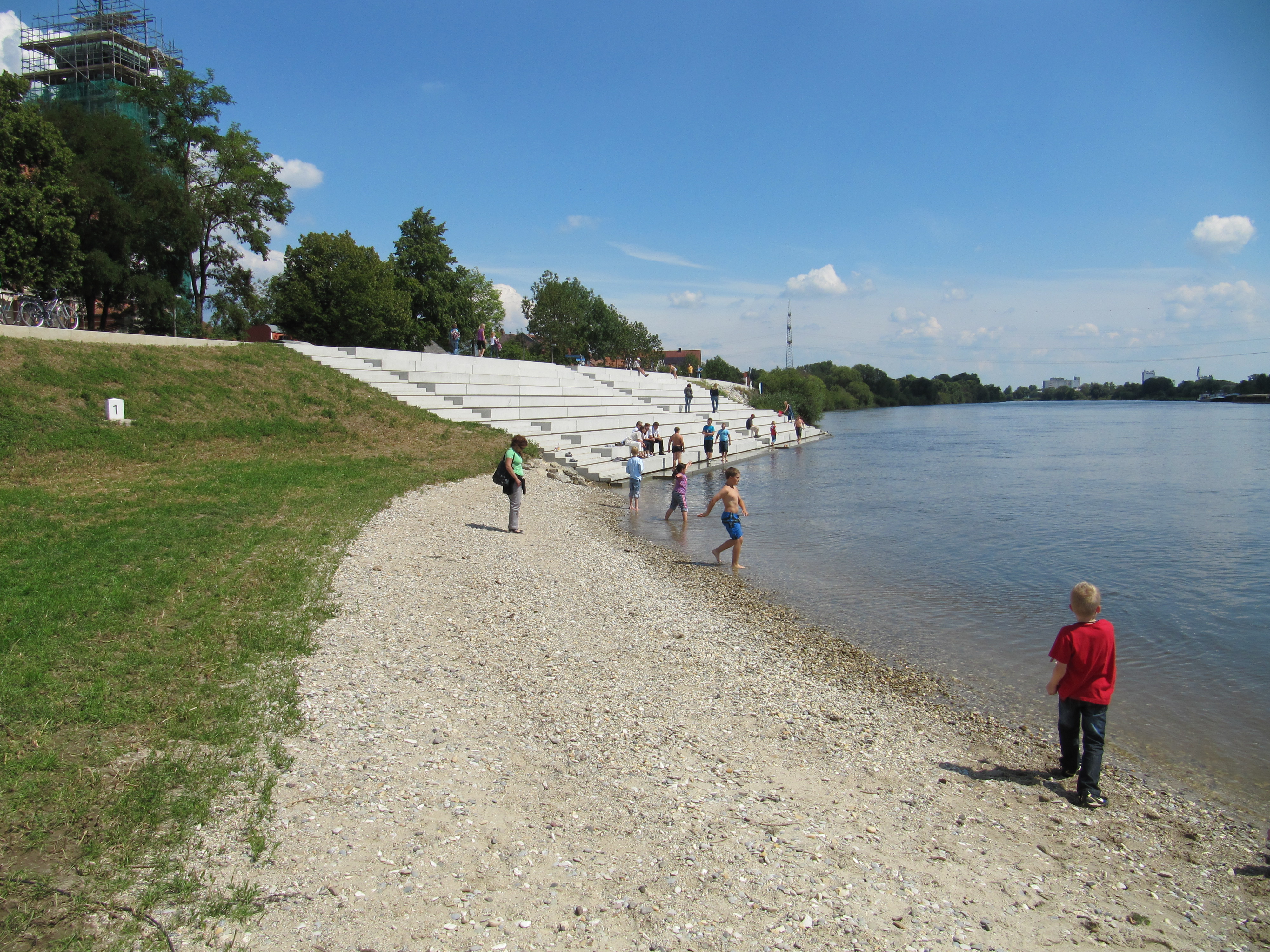 Im Vordergrund rechts Kinder an der Donau, in der Mitte der Kiesstrand und links eine Wiese mit Böschung zur im Hintergrund zu sehende Kirche mit eingerüstetem Turm, rechts davon die Treppenanlage mit einigen Personen