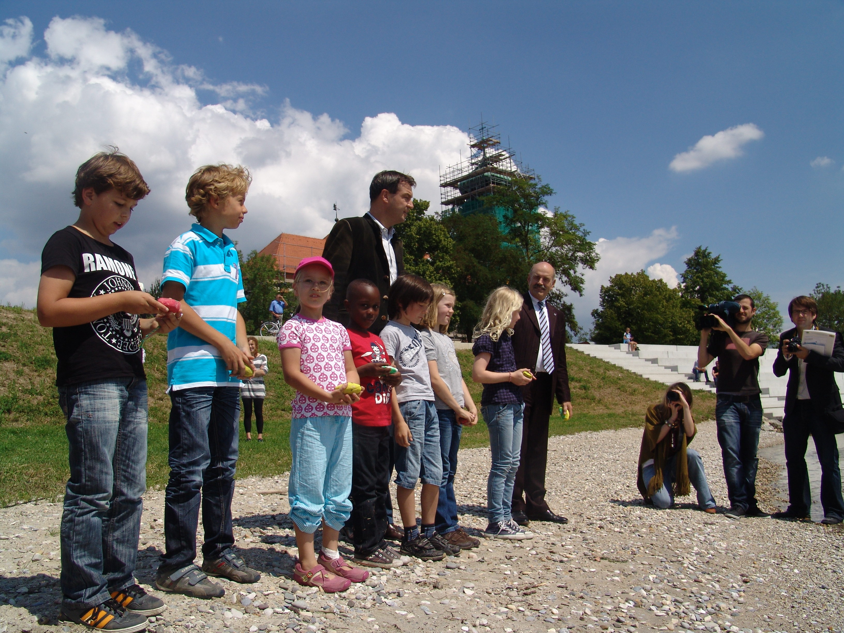 Minister Söder und Oberbürgermeister Schaidinger stehen mit sieben Grundschulkindern am Kieselstrand und halten bemalte Steine in den Händen, rechts davon drei Reporter, im Hintergrund die Treppenanlage und die Kirche mit eingerüstetem Turm