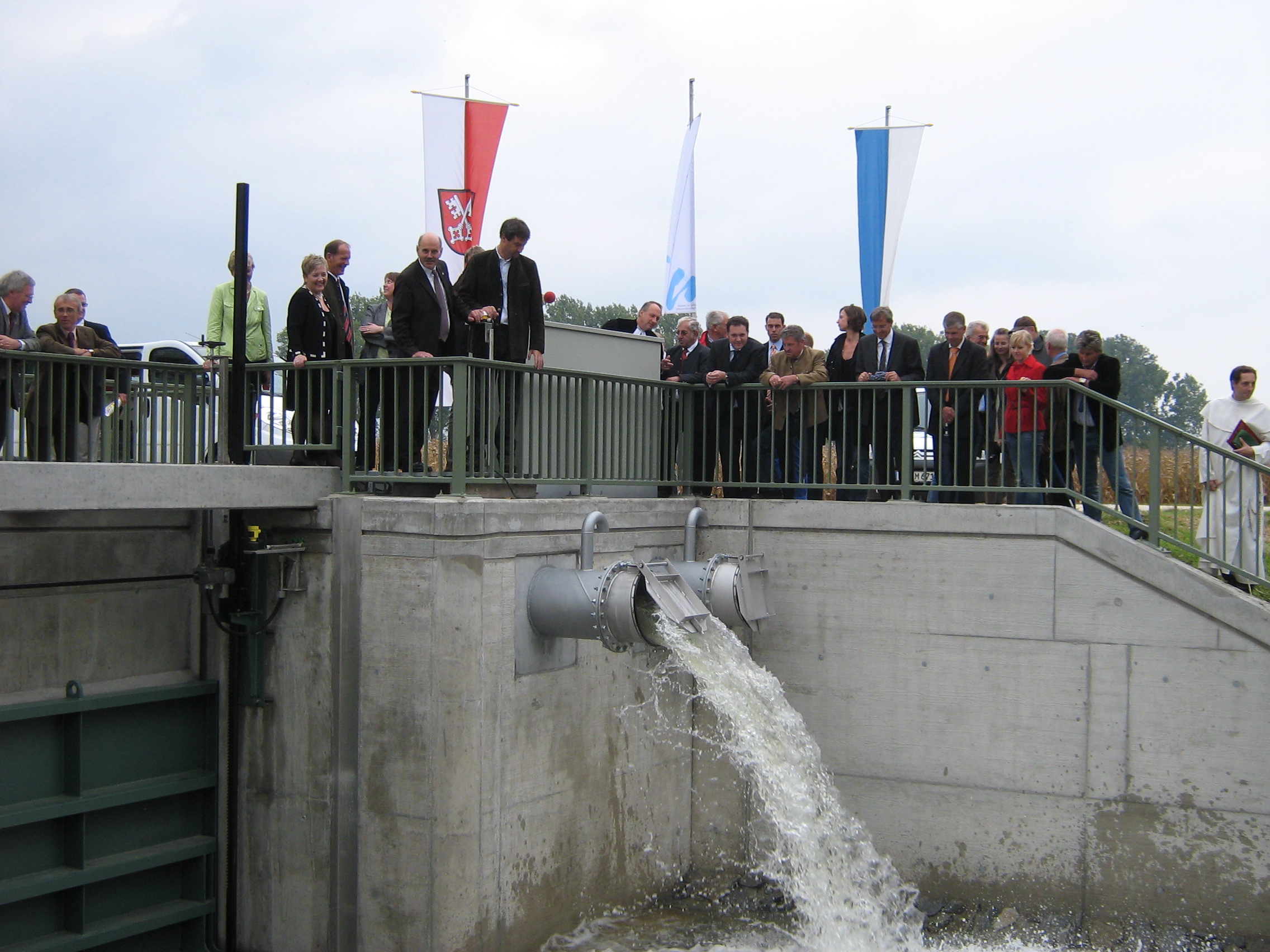 Eine Personengruppe steht verteilt entlang des Geländers am Übergangs des Schöpfwerkes. Darunter sind die Rohrschächte und die geschlossene Schütztafel zu sehen. Aus dem linken Rohrschacht strömt Wasser. Im Hintergrund sind drei Fahnen und Fahrzeuge zu erkennen.