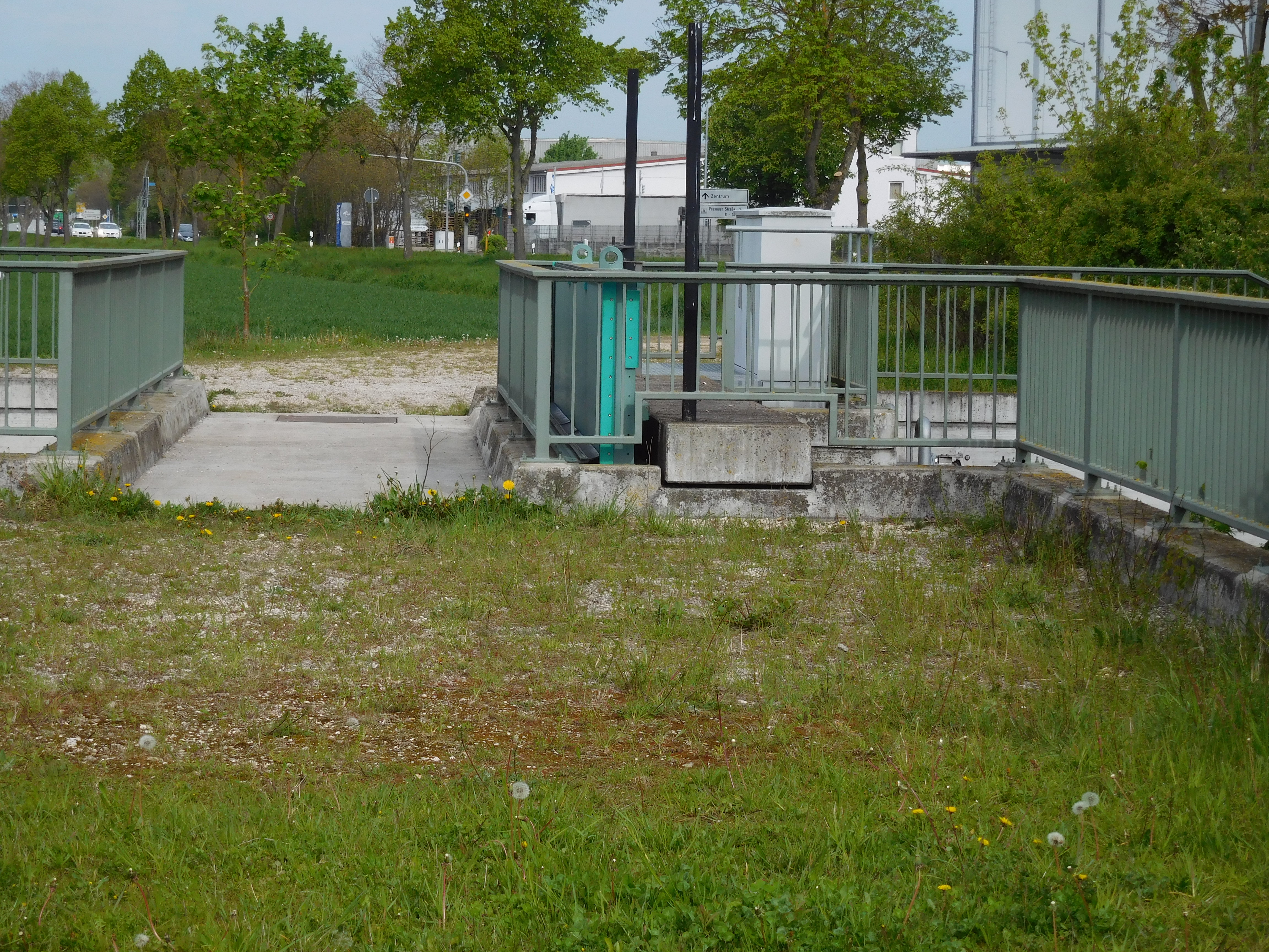 Längs im Bild der Übergang des Schöpfwerks, mit beidseitigem grünen Geländer. Im Hintergrund ein Feld, die Straße und Industriebebauung.