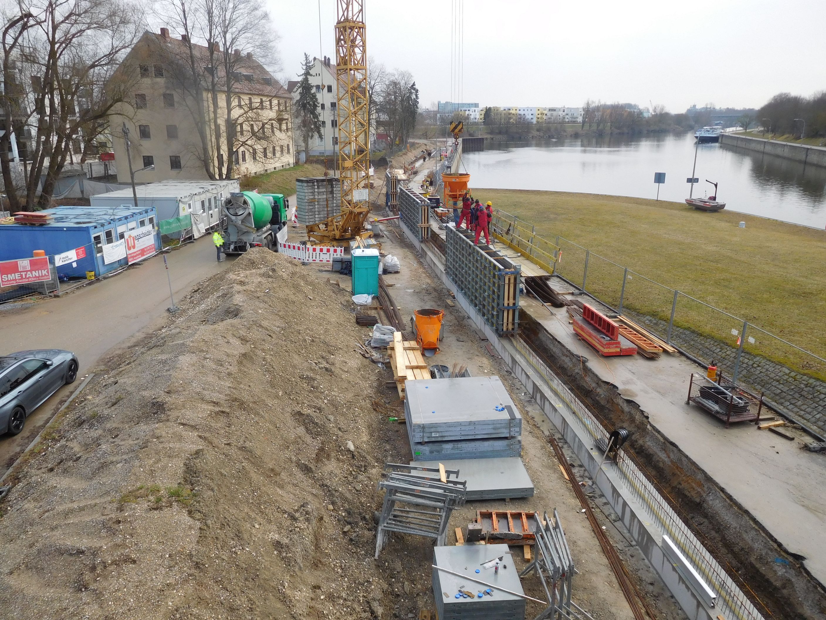 Überblick auf die Baustelle, links Bebauung, Baucontainer und ein Erdhaufen, rechts die Schalungsbretter, die Hochwasserschutzmauer und die Donau, im Hintergrund ein Kran
