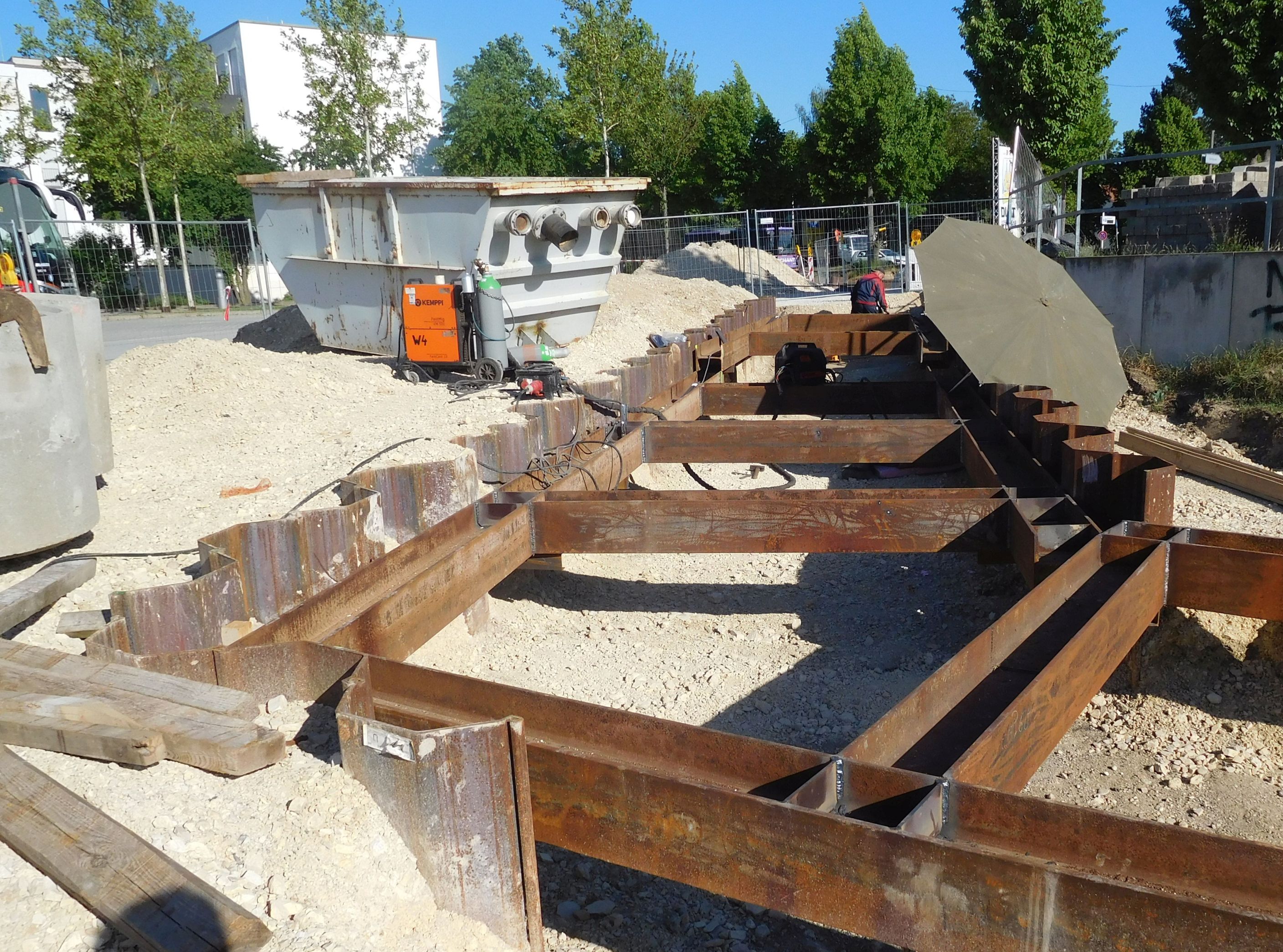 Zu sehen ist ein Stahlrahmen zum Aussteifen der eingebrachten Spundwände, die rings um den Rahmen zu sehen sind. Links ein Bauschuttcontainer und Bauutensilien, im Hintergrund Bebauung und Bäume