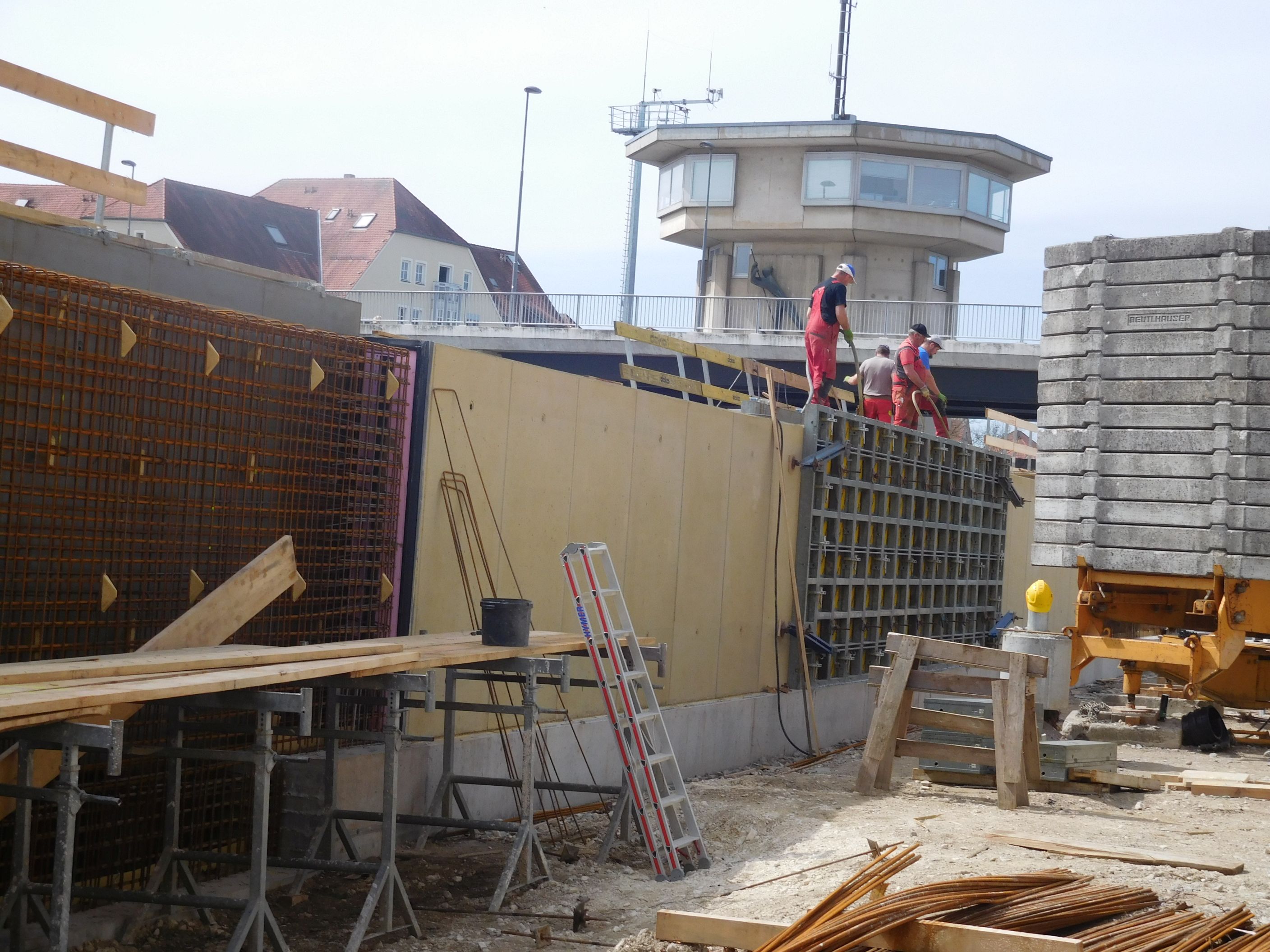 Quer durch das Bild die Schalung für die Hochwasserschutzmauer, vier Arbeiter sind auf der Mauer, rechts daneben das Gegengewicht eines Krans, im Hintergrund die Kanalbrücke mit Schleusenhaus