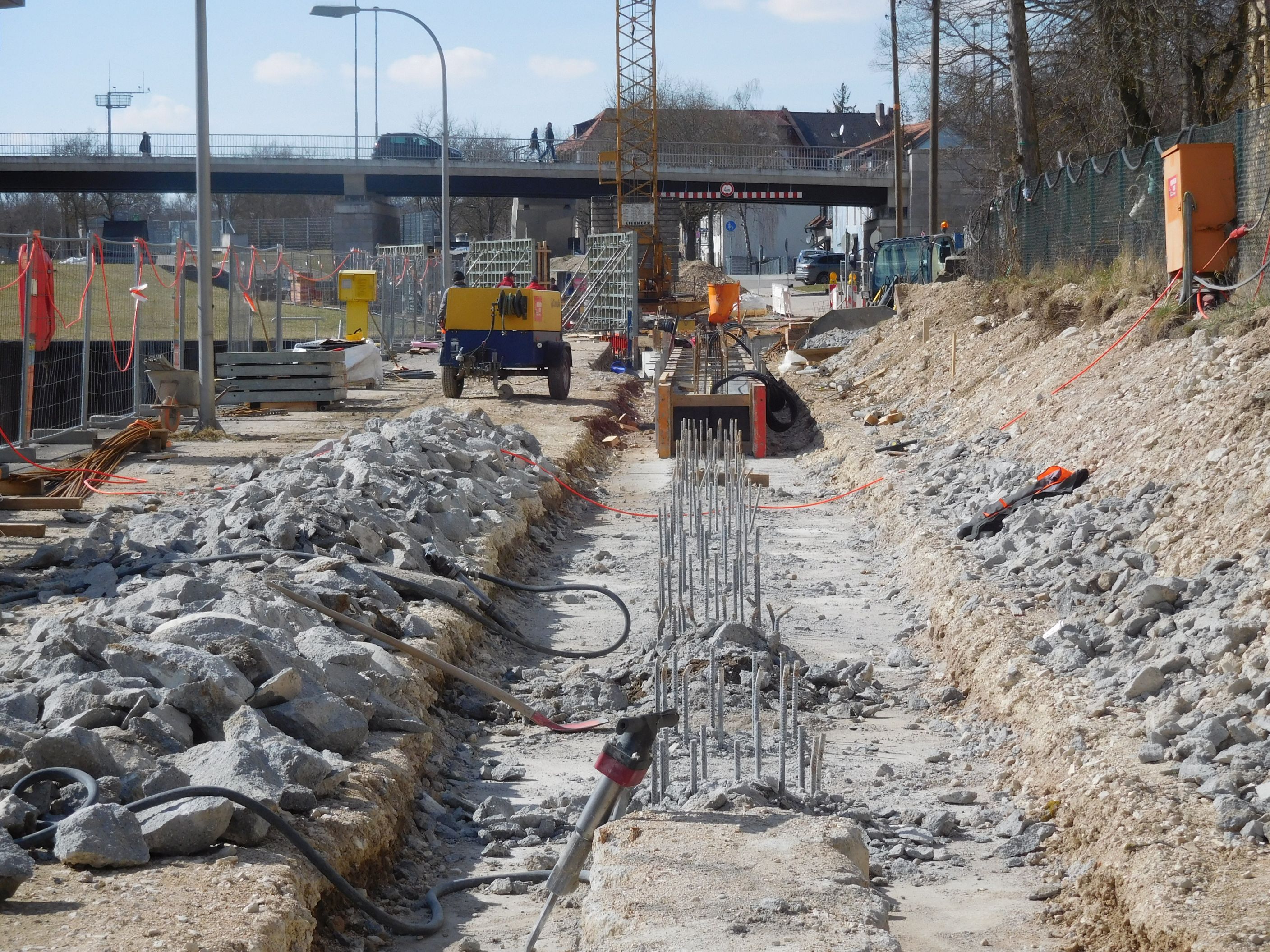 In der Mitte des Bildes nach hinten verlaufend ist die Bewehrung der Bohrpfahlwand zu sehen. An beiden Seiten liegten die abgetrennten, überschüssigen Betonstücke. Im Hintergrund ein Kompressor, ein Baukran, die querende Kanalbrücke und die Bebauung. Einzelne Werkzeuge sind zu sehen.