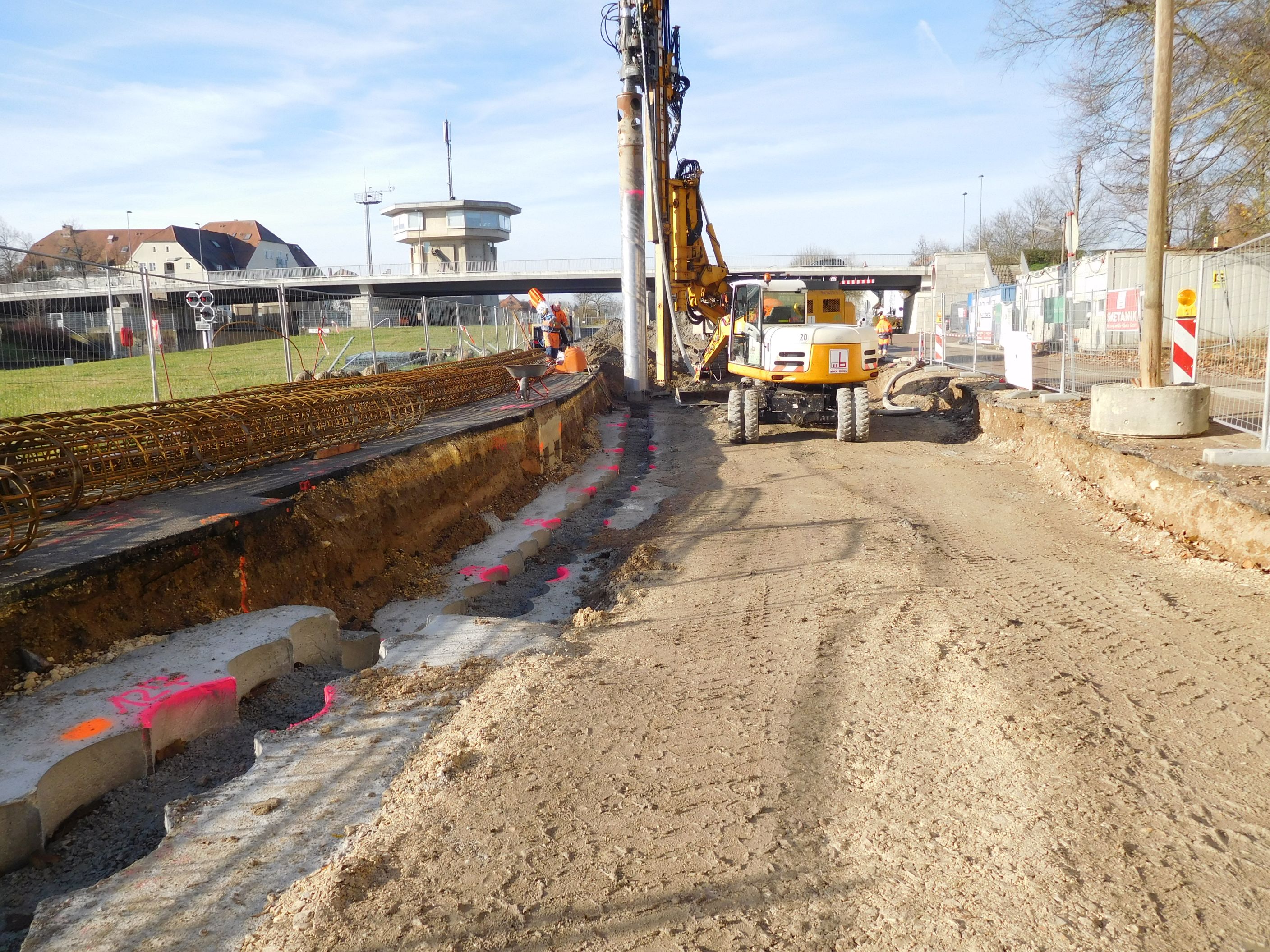  Rechts im Bild die Baustrasse, dahinter ein Bohrgerät und die Kanalbrücke, rechts steht ein Bauzaun, links entlang der Baustraße liegen die Bewehrungskörbe und ein Bauzaun steht dahinter.