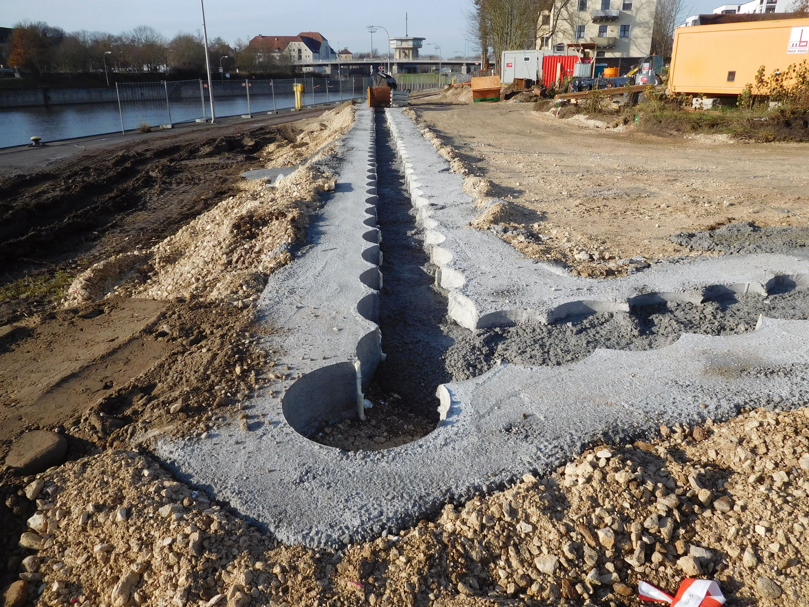 In der Mitte des Bildes nach hinten und zur rechten Seite verlaufend die Bohrschablone aus Beton, rechts die Baustrasse, links die Baustrasse und der Schleusenkanal. Im Hintergrund Baucontainer, Bebauung und die querende Kanalbrücke mit Schleusenhaus.