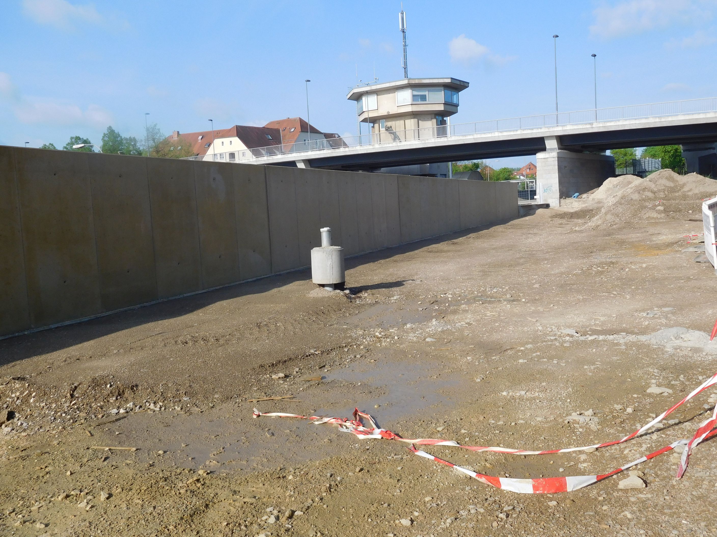 Das geräumte Baufeld mit einem Beweissicherungspegel in der Mitte des Bildes, im Hintergrund die Hochwasserschutzmauer und die querende Kanalbrücke mit Schleusenhaus. Ein Absperrband liegt im Vordergrund.