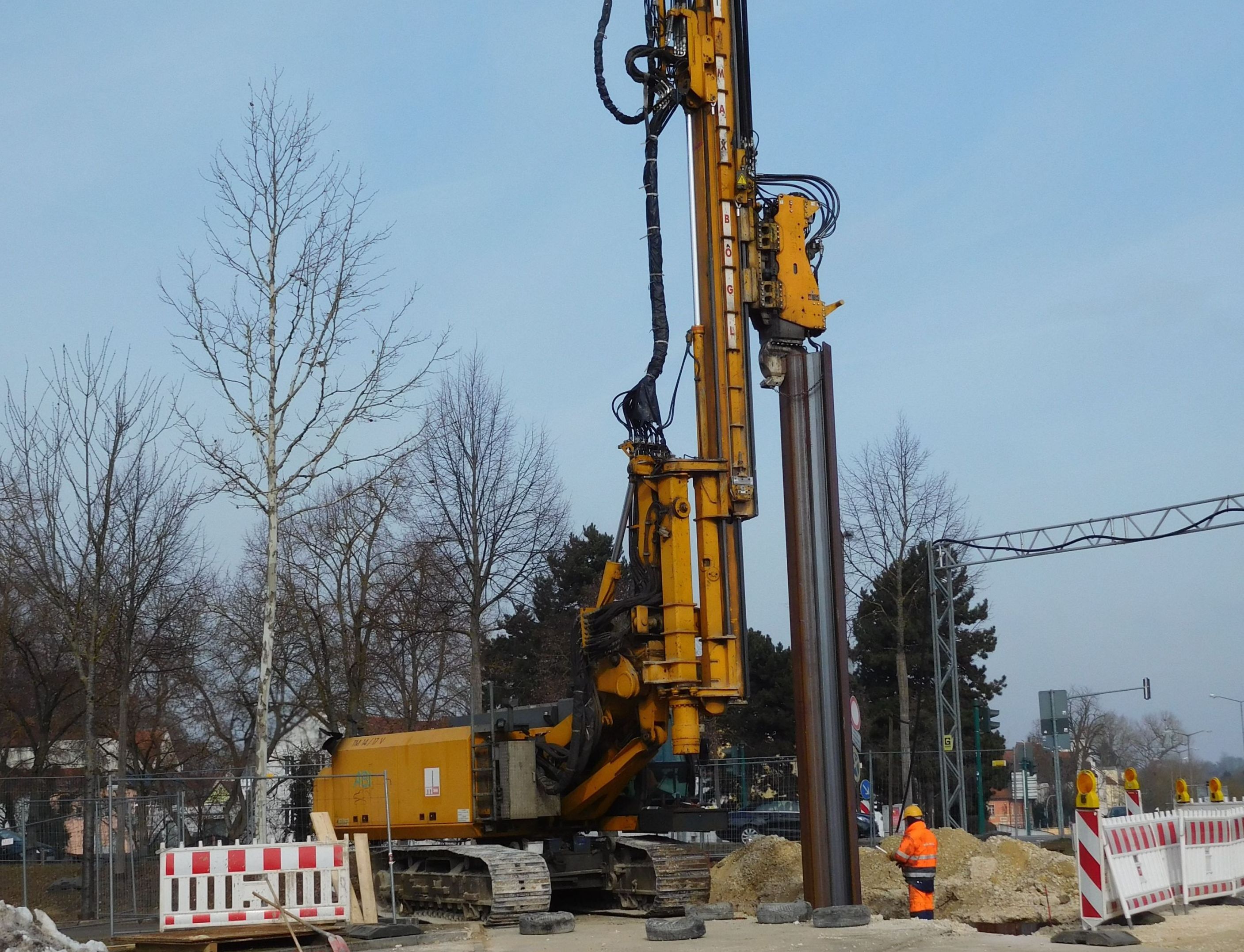 In der Mitte des Bildes bringt eine Baumaschine eine Spundwand in den Boden ein. Ein Bauarbeiter steht neben den Spundwand. Links Bäume und eine Bake, rechts stehen Absperrbaken und eine Überleitung für das Stromkabel, dahinter ist eine Straßenkreuzung zu erkennen.