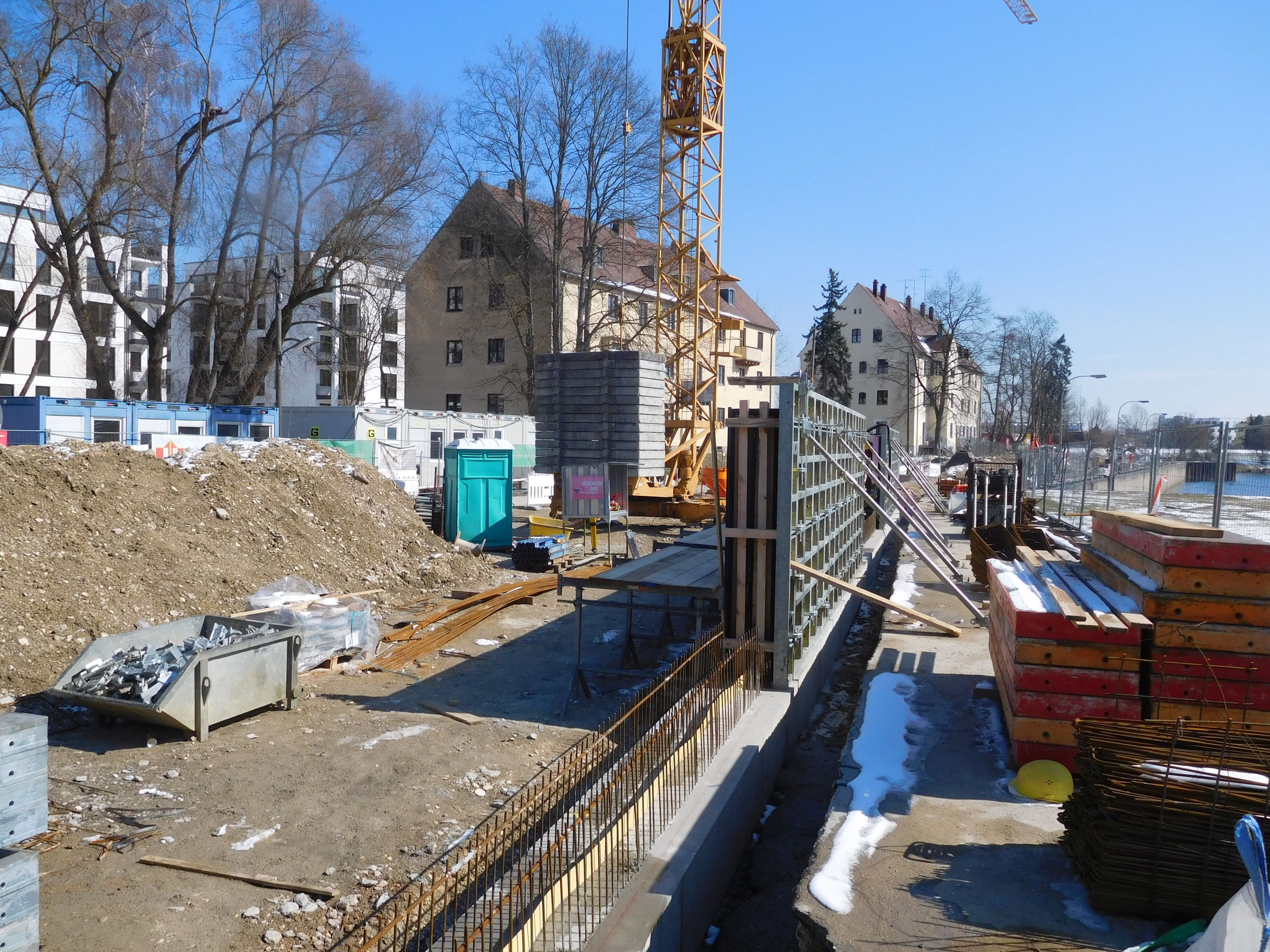 In der Mitte des Bildes nach hinten verlaufend die Schalung für die Hochwasserschutzmauer, links davon Baucontainer, Bebauung und Bauutensilien, rechts Schalungsbretter, ein Kran steht im Hintergrund