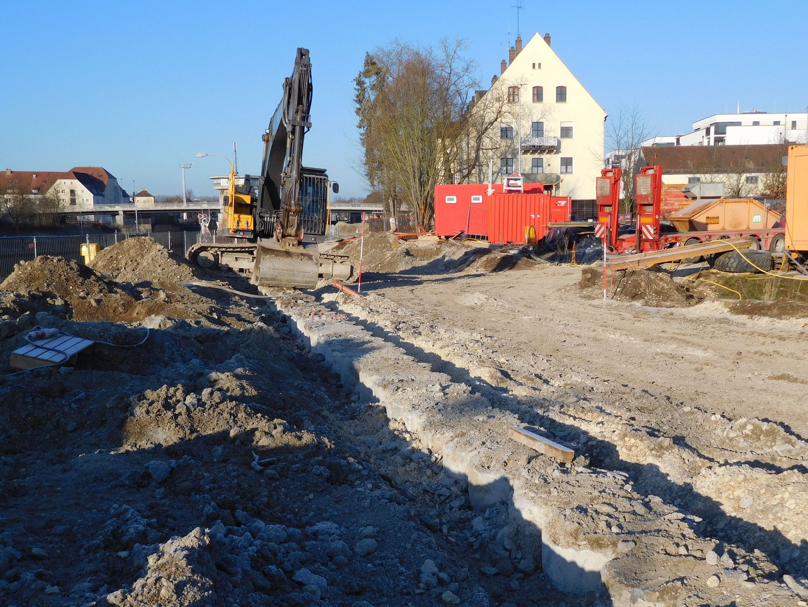 Diagonal nach hinten verlaufend die Bewehrungspfähle. Ein Bagger steht auf den Pfählen und nimmt das Material der Bohrschablone mit der Schaufel weg. Links davon Erdreich, dahinter die Kanalbrücke, rechts die Baustelleneinrichtung und Bebauung. 