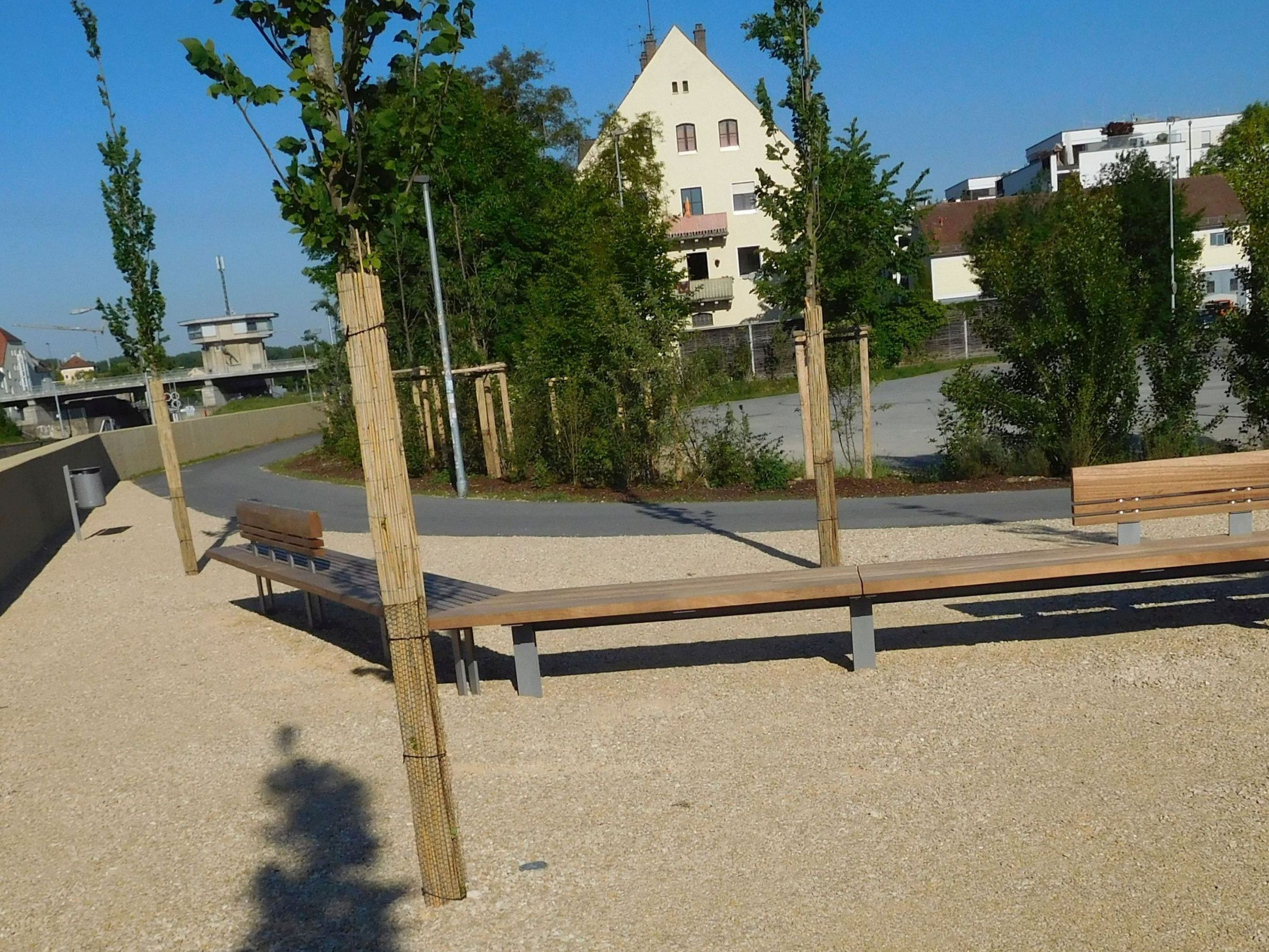 Die Ecke der Mündung des Flusses Regen in die Donau mit neu angelegtem, asphaltierten Radweg, einer Sitzbank in L Form aus Holz und neuer Bepflanzung bei blauem Himmel, Häuser im Hintergrund