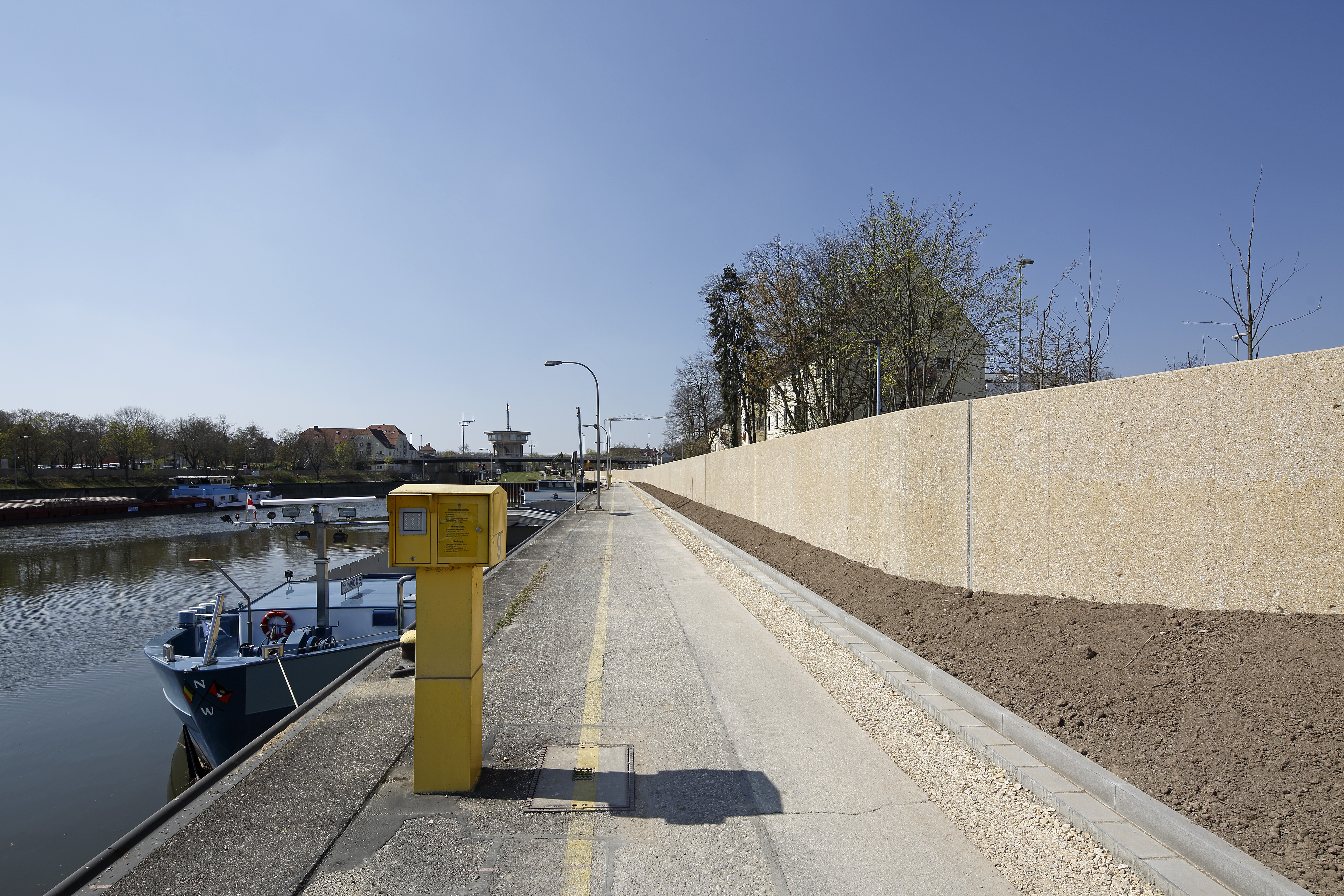 Rechts im Bild verläuft die Hochwasserschutzwand, dahinter eine Wohnblock. In der Mitte der Uferweg mit Laternen und einer gelben Anschlusssäule. Links die Donau mit ein paar kleine Booten. Im Hintergrund quert die Schleusenbrücke und das Schleusenhaus.