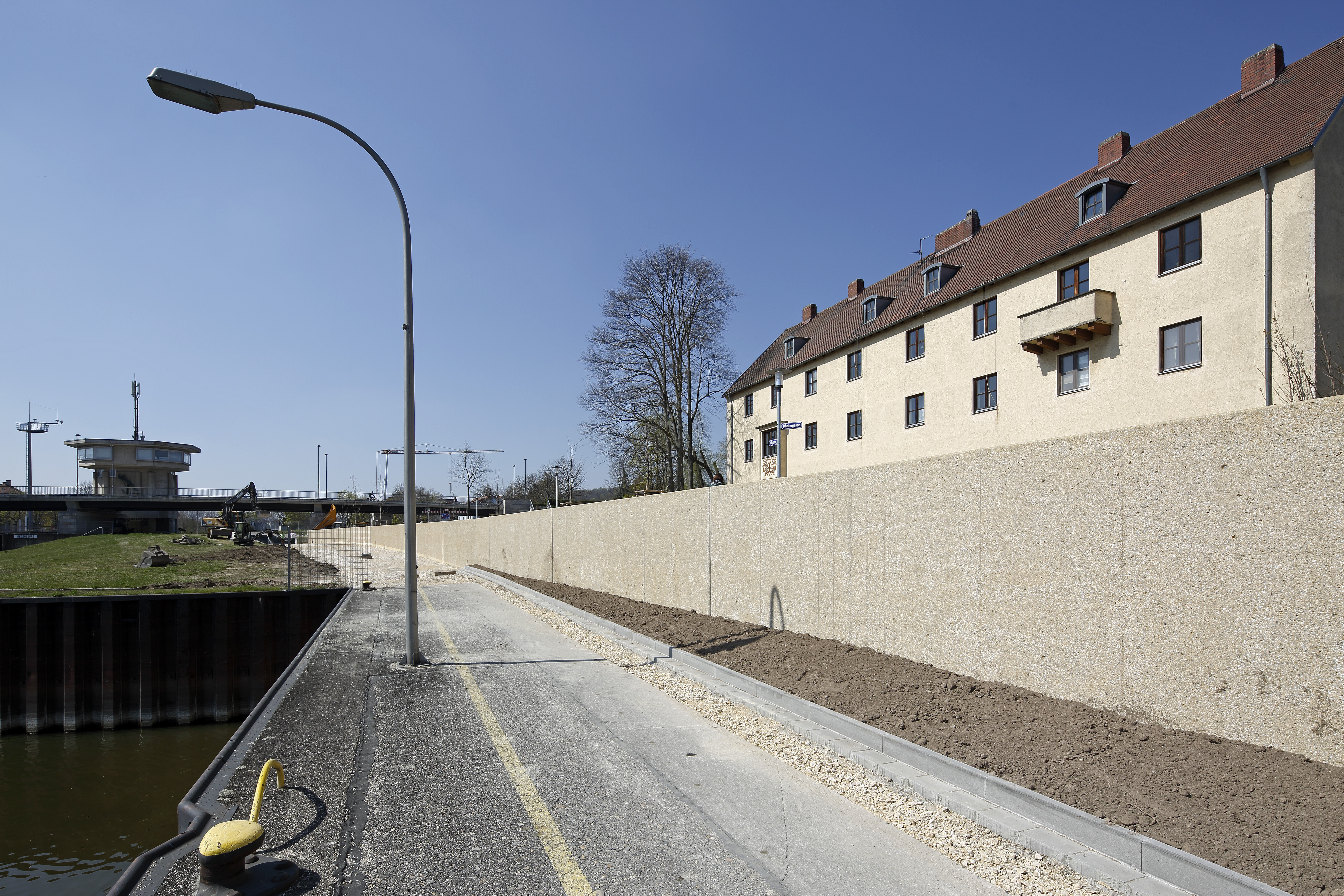 Rechts im Bild verläuft die Hochwasserschutzwand, dahinter eine Wohnblock. In der Mitte der Uferweg mit einer Laterne, links die Spundwand zur Einfahrt in die Schleuse. Im Hintergrund quert die Schleusenbrücke und das Schleusenhaus.