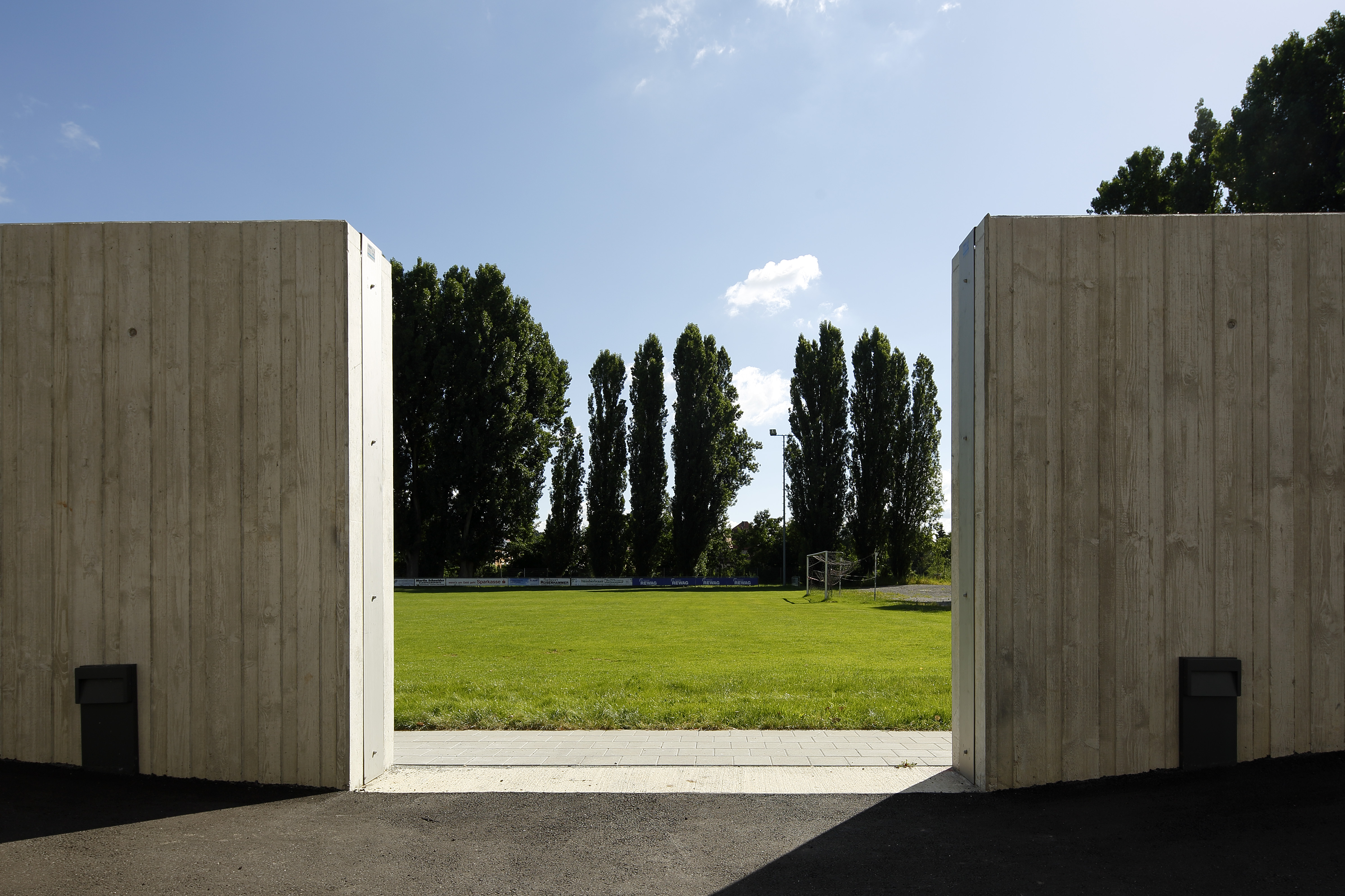 Auf beiden Seiten des Bildes die betonierte Hochwasserschutzwand, in der Mitte die Maueröffnung für mobilen Lückenschluss zum Sportplatz. Im Hintergrund quert die Bandenwerbung und eine Baumreihe bei strahlend blauem Himmel.