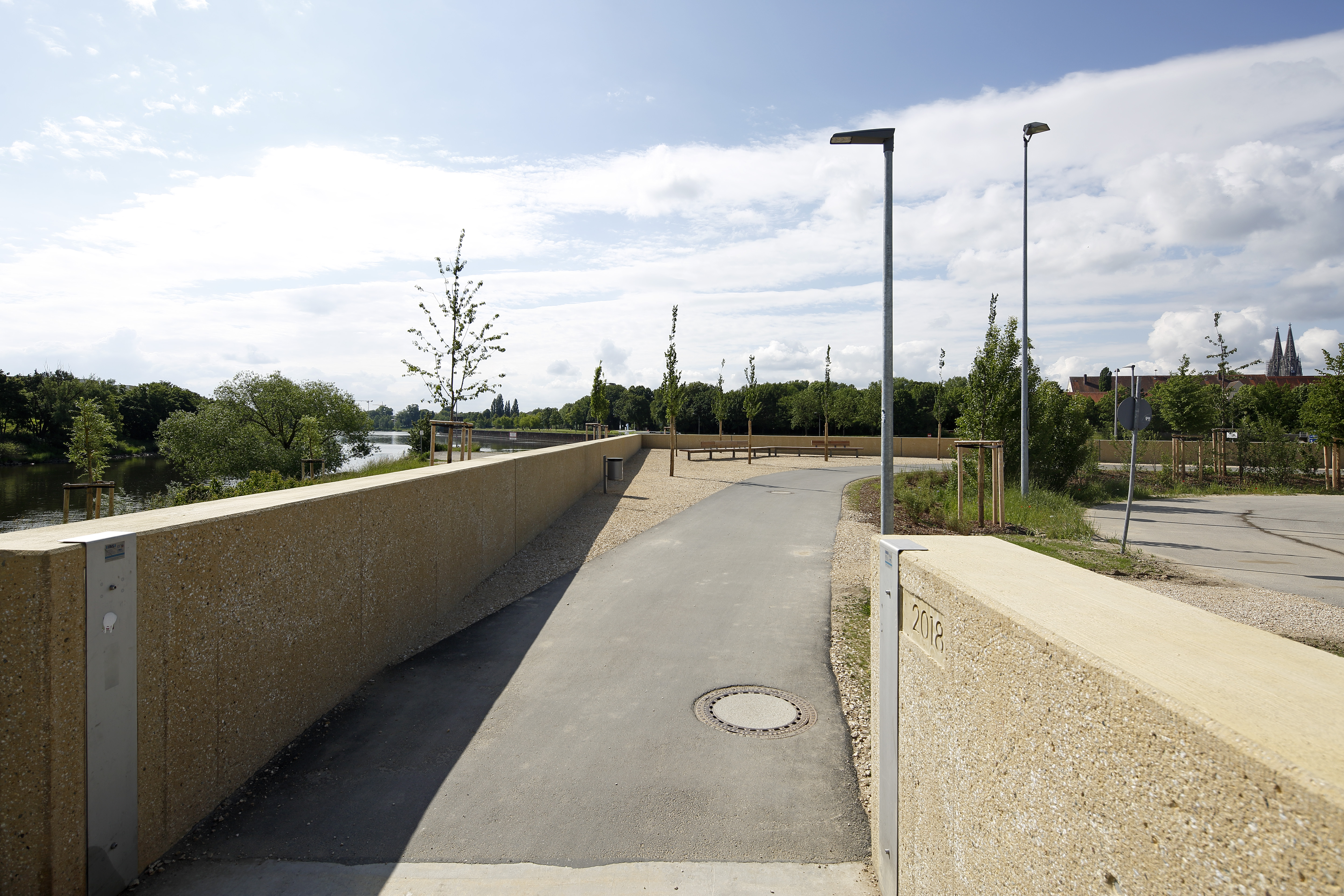 Blickrichtung Regenmündung, ganz links im Bild ist noch der Fluß Regen zu erkennen, daneben die HWS Mauer, in der Mitte die Durchfahrt für den Radweg. Rechts endet die HWS Mauer. Hier wurde als Baujahr die Zahl 2018 eingebracht. Im HW Fall wird hier ein mobiler Lückenschluss eingebracht, im Hintergrund die abknickende HWS Mauer und die hölzerne Sitzbank, bei leicht bewölktem Himmel