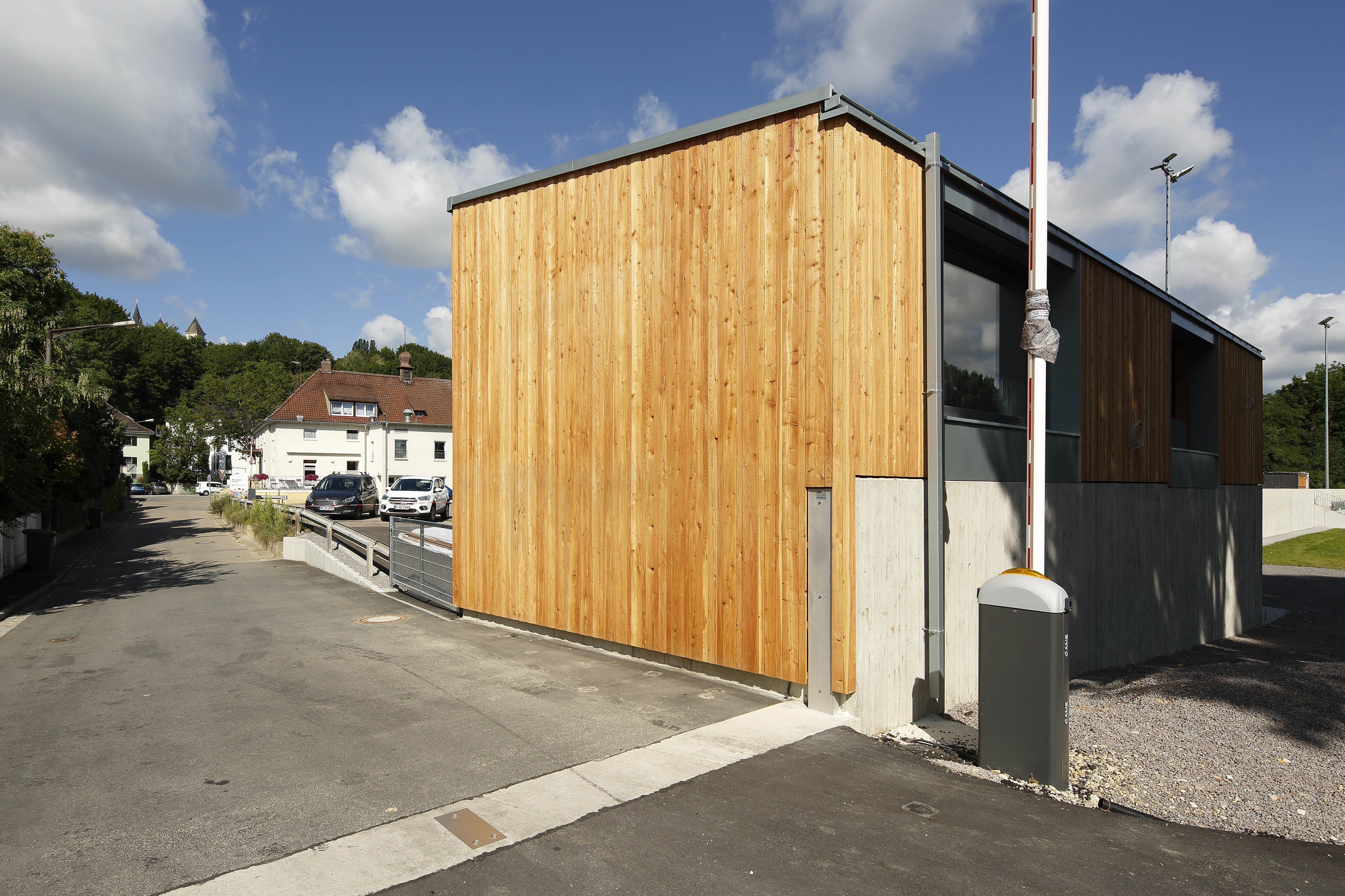 Neu errichtetes Nebengebäude der SG Walhalla mit Holzfassade, links die Durchfahrt mit mobilem Lückenschluss, im Hintergrund der Parkplatz und das Vereinsgebäude der SG Walhalla.