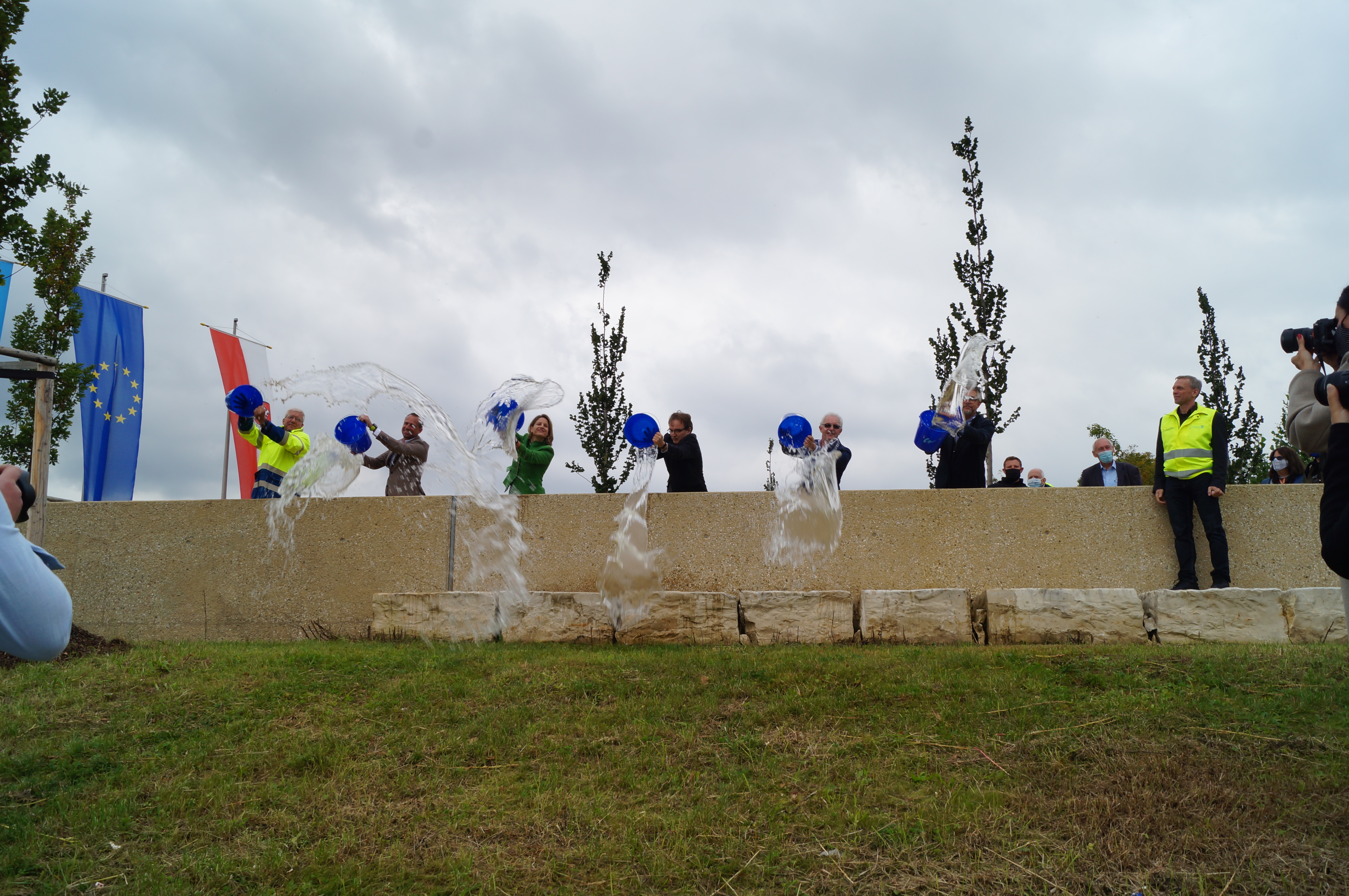 Quer im Bild verläuft die Hochwasserschutzwand, dahinter stehen die Verantwortlichen und schütten aus blauen Eimern Wasser über die Hochwasserschutzwand. Links im Bild hinter der Mauer die Fahnen der Stadt Regensburg und der EU bei bewölktem Himmel. 