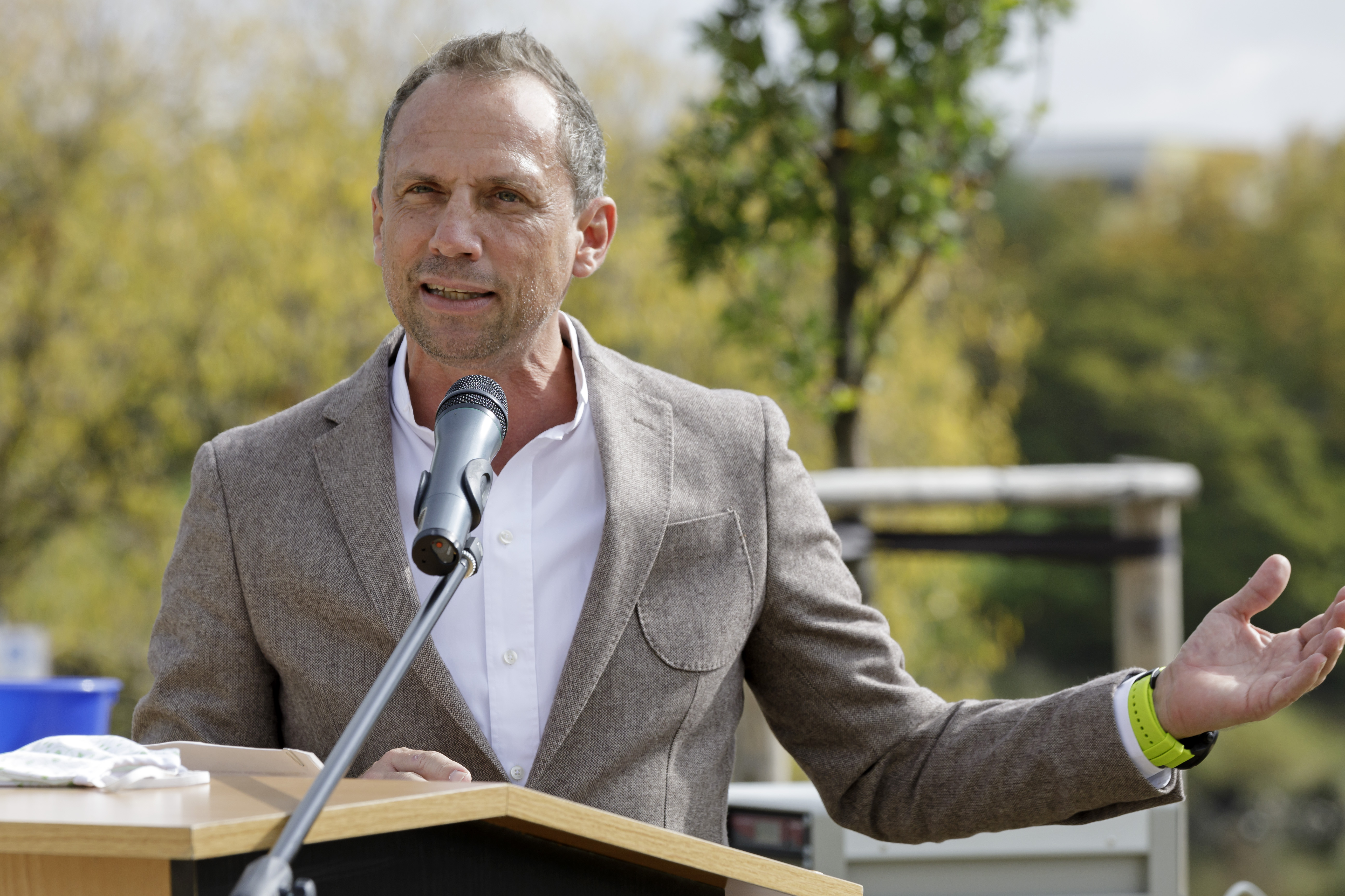 Minister Glauber steht hinter dem Mokrofon am Rednerpult. Sein linker Arm ist seitlich ausgestreckt. Im Hintergrund Bepflanzung. 