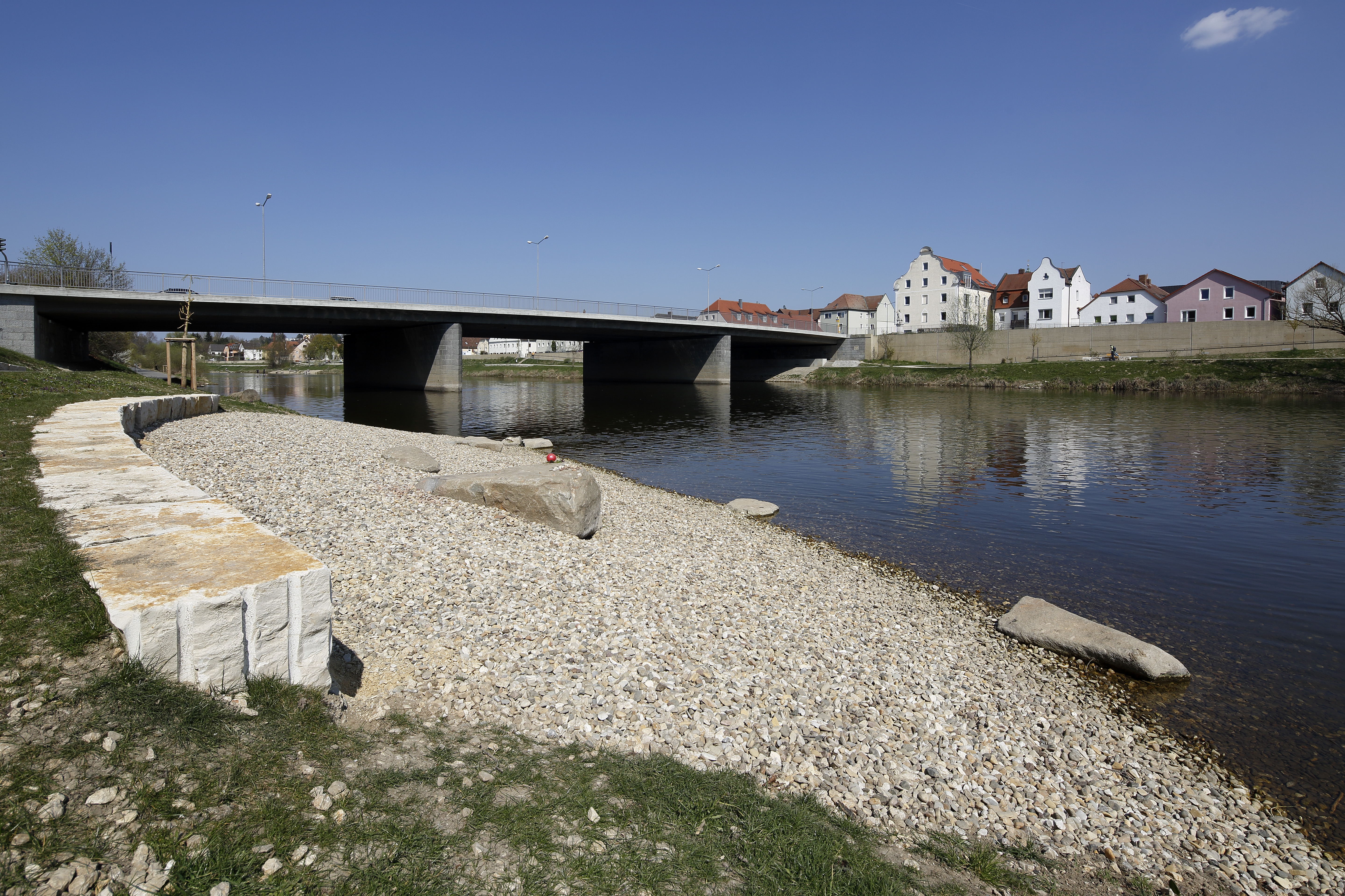Rechts der Fluß Regen, darüber die Reinhausener Brücke und einer Häuserreihe am rechten Ufer, in Bildmitte eine Kiesbank mit bogenförmig aneinandergereihten Steinquadern zum Sitzen bei strahlend blauem Himmel