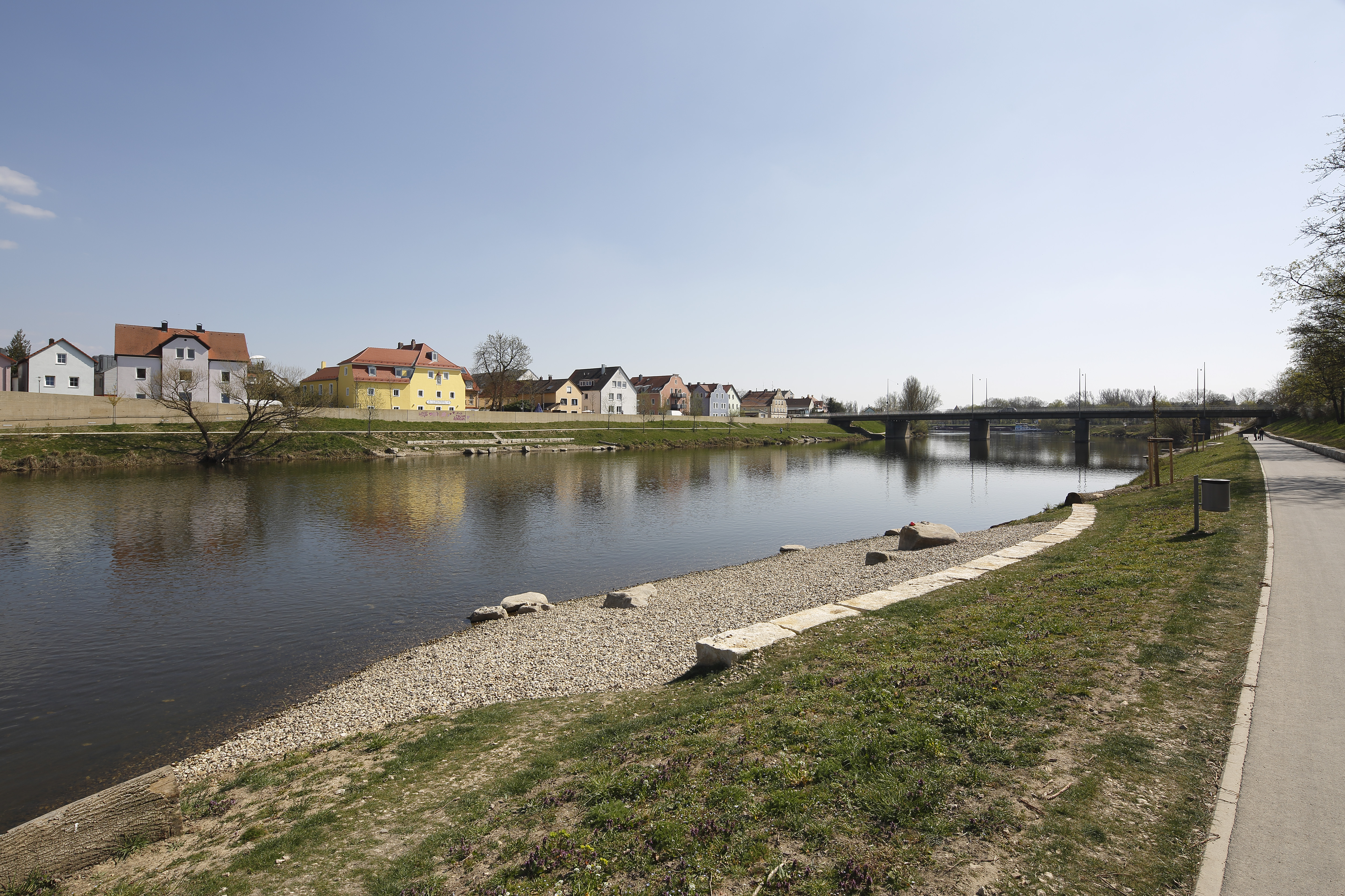 In Bildmitte der Fluß Regen, im Hintergrund am anderen Ufer eine bunte Häuserreihe mit grüner Böschung und die Frankenbrücke, im vorderen Bereich eine Kiesbank mit einer Reihe Steinquader zum Sitzen, große Steine liegen auf der Kiesbank und im Wasser